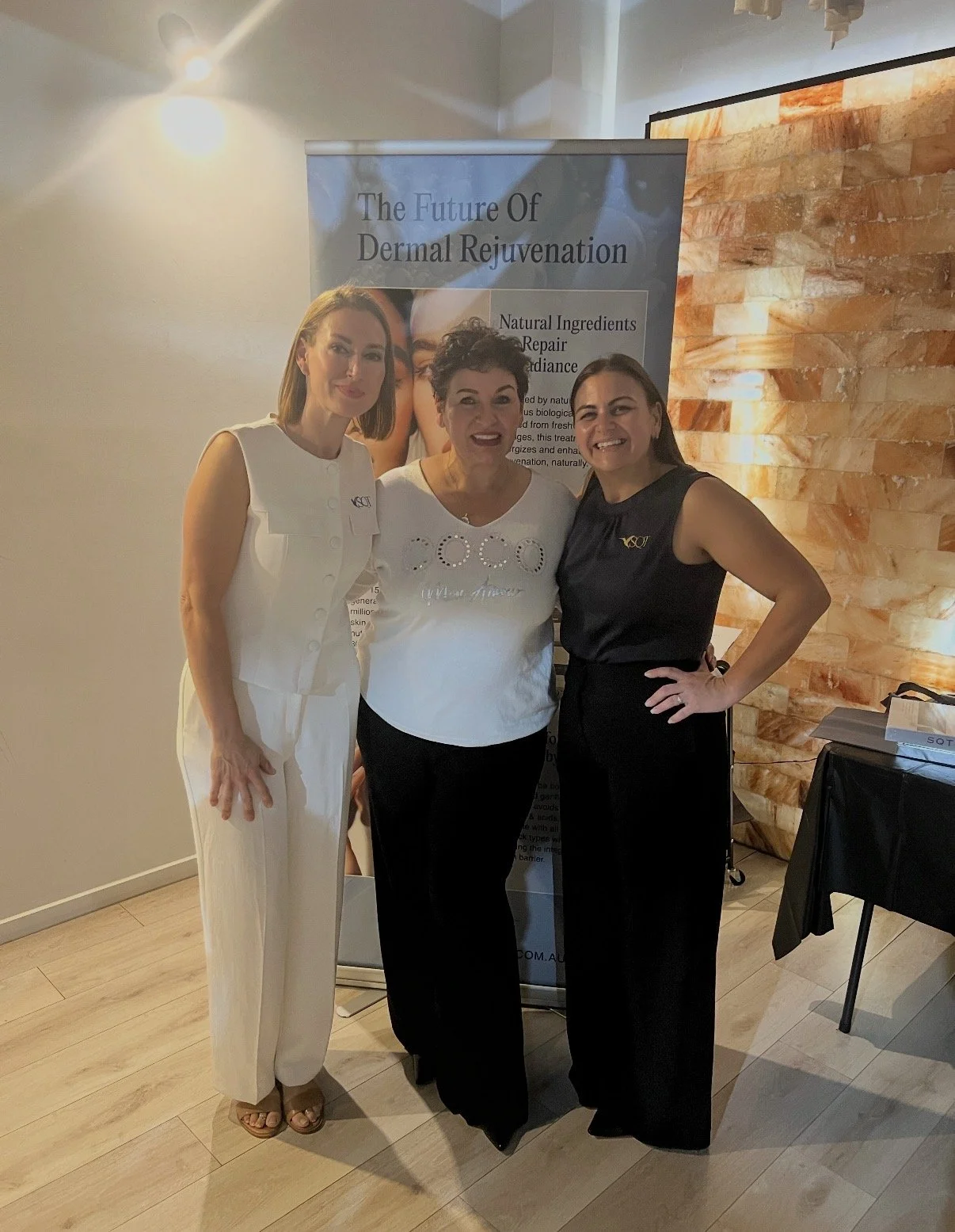 Three women standing together indoors, smiling, with a promotional banner behind them that reads "The Future Of Dermal Rejuvenation" and mentions natural ingredients, repair, and radiance.