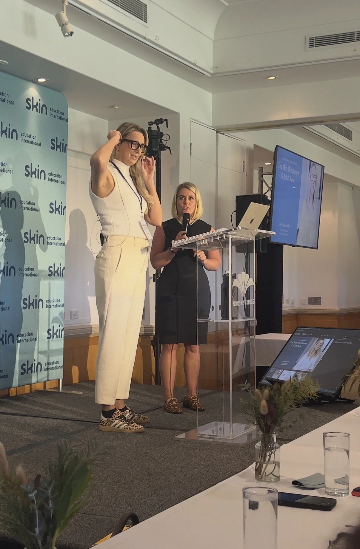 Two women standing at a podium during a presentation at a conference, with one woman speaking into a microphone and the other adjusting her glasses. There is a large screen displaying a presentation slide and a backdrop with the logo 'skin education 