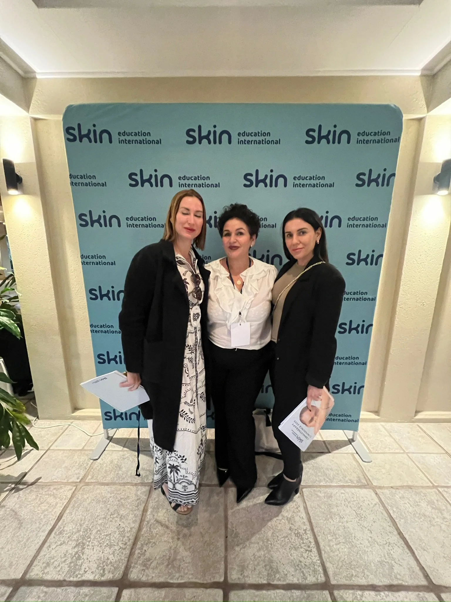 Three women standing in front of a backdrop with 'skin education international' logos, holding papers, at an indoor event.
