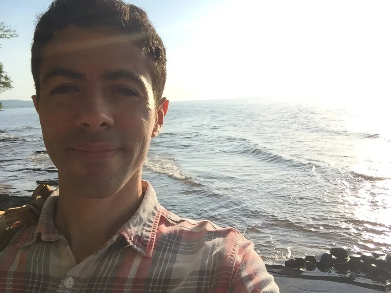 A young man taking a selfie by the water, with the ocean and the horizon in the background during the daytime.