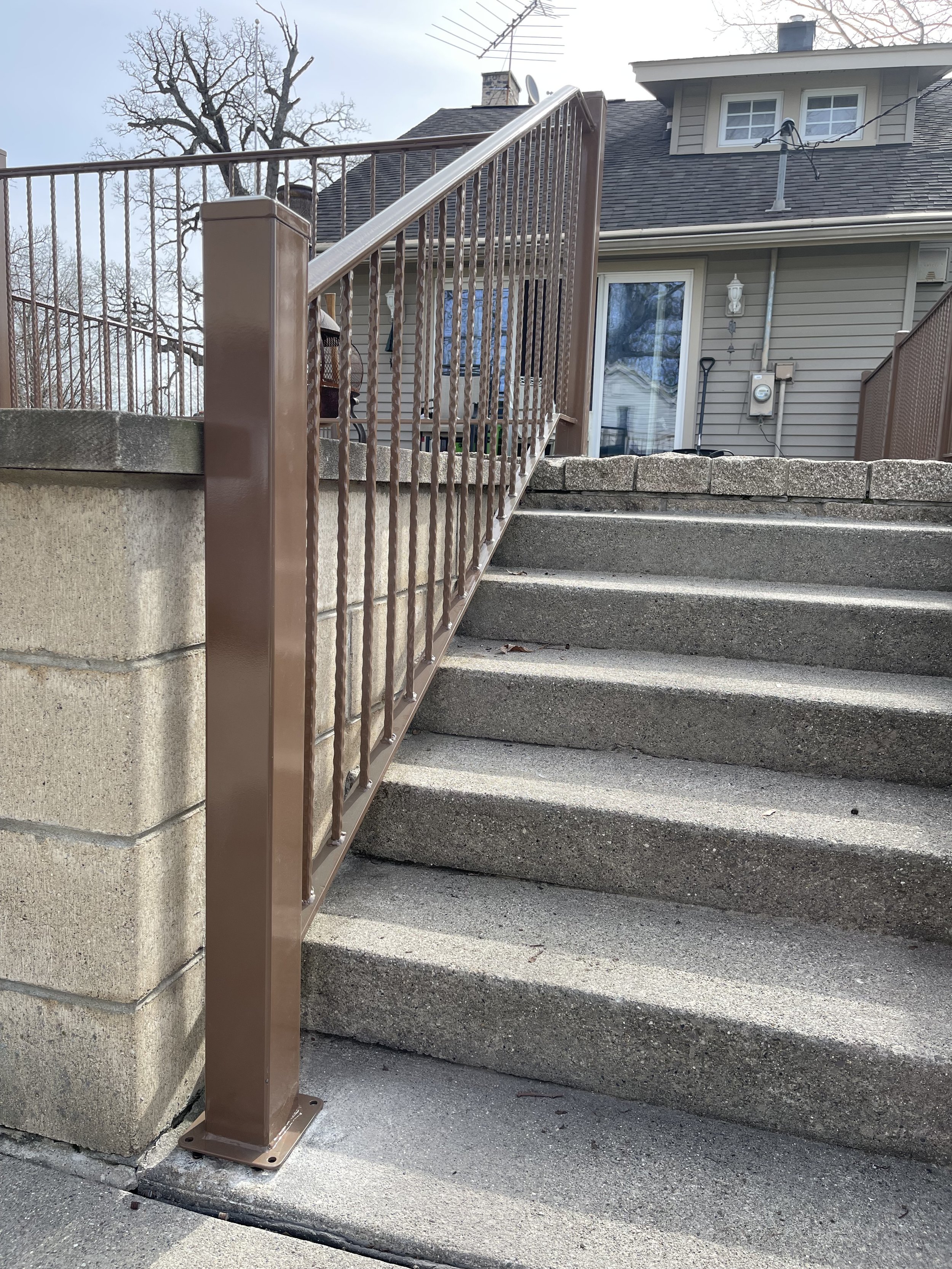 Concrete stairs with a metal railing leading up to a house with beige siding, a sliding glass door, and outdoor lighting. A tree is visible in the background.