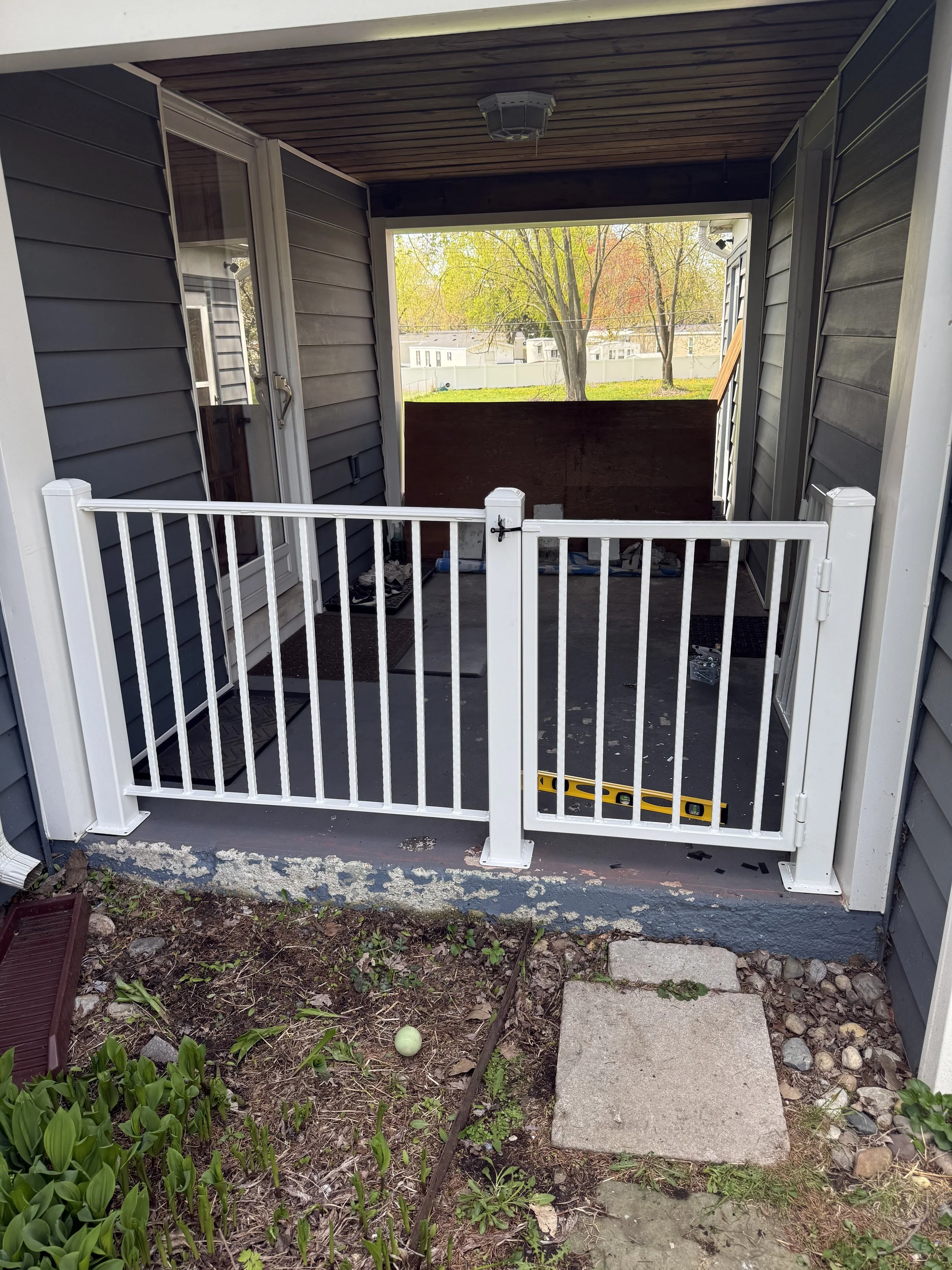 A small porch with a white safety gate and a concrete stepping stone, leading to a backyard with trees, grass, and a white fence, during fall.