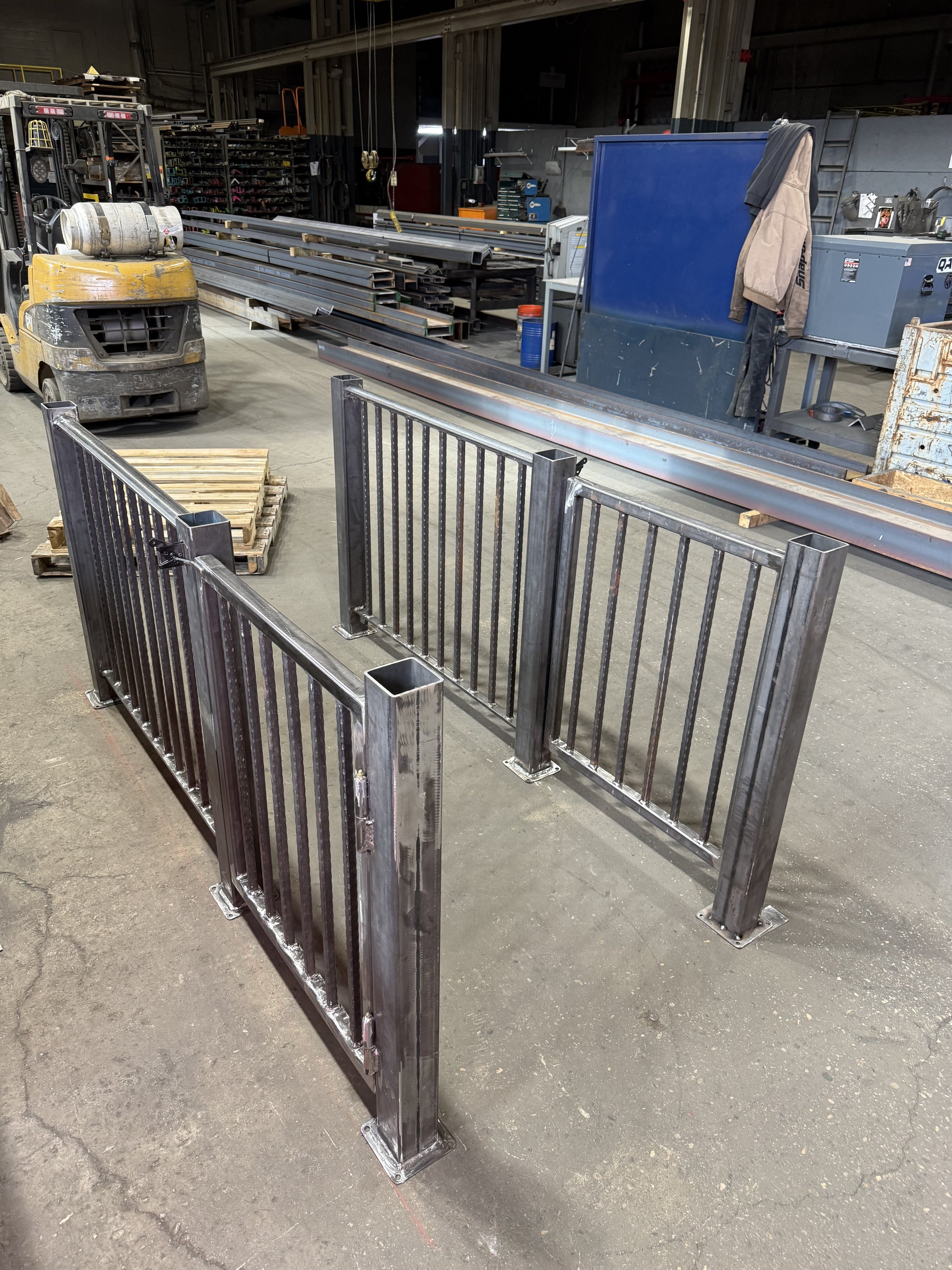 In a warehouse or workshop, metal security fences are being assembled, with metal fence panels and a forklift nearby. There are various tools, equipment, and shelving in the background.