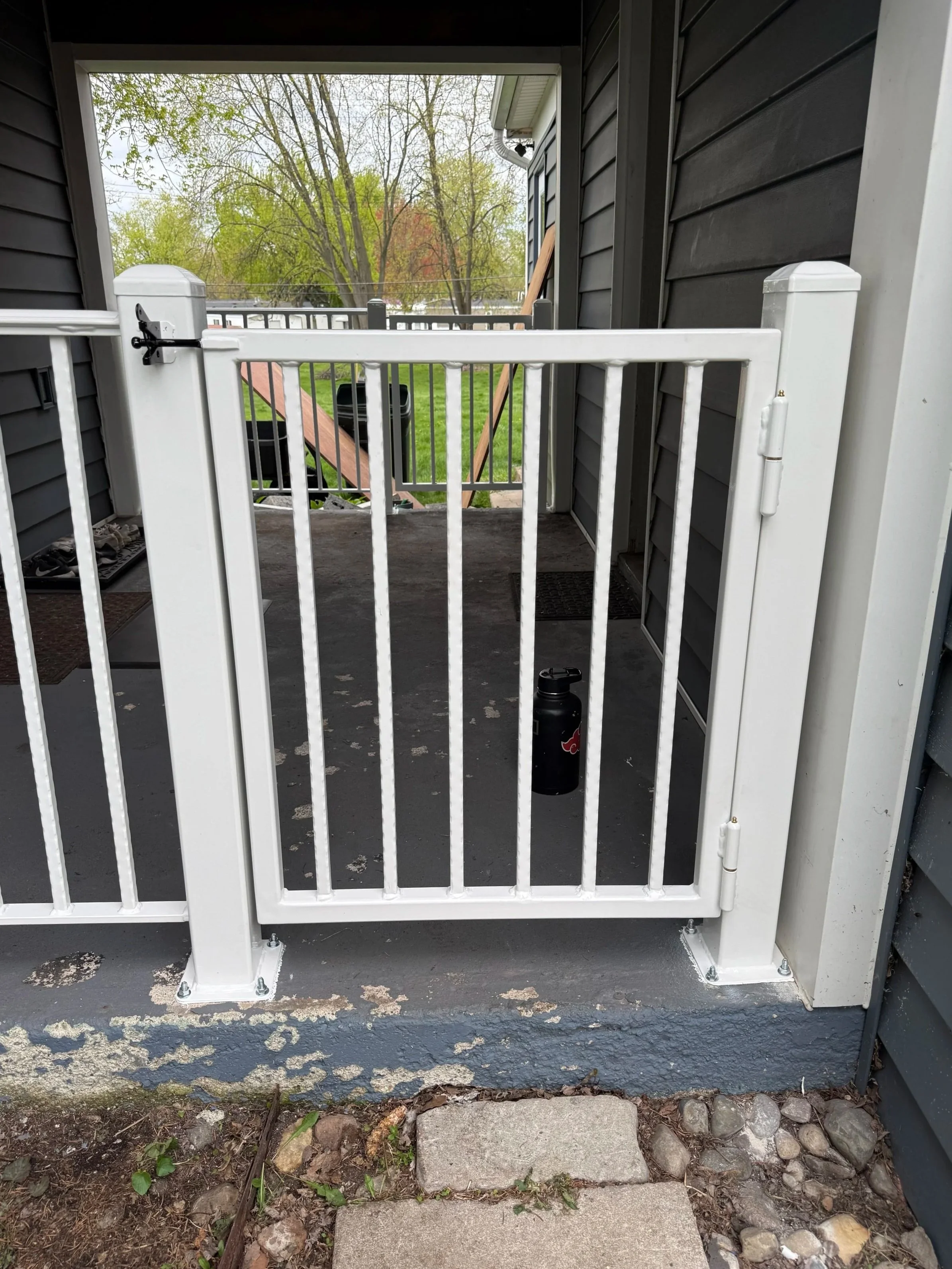 A small child safety gate mounted on the front porch of a house, with a black water bottle on the porch floor in front of it. The porch has black siding, and beyond the gate, there is a wooden railing and a backyard with trees and grass.