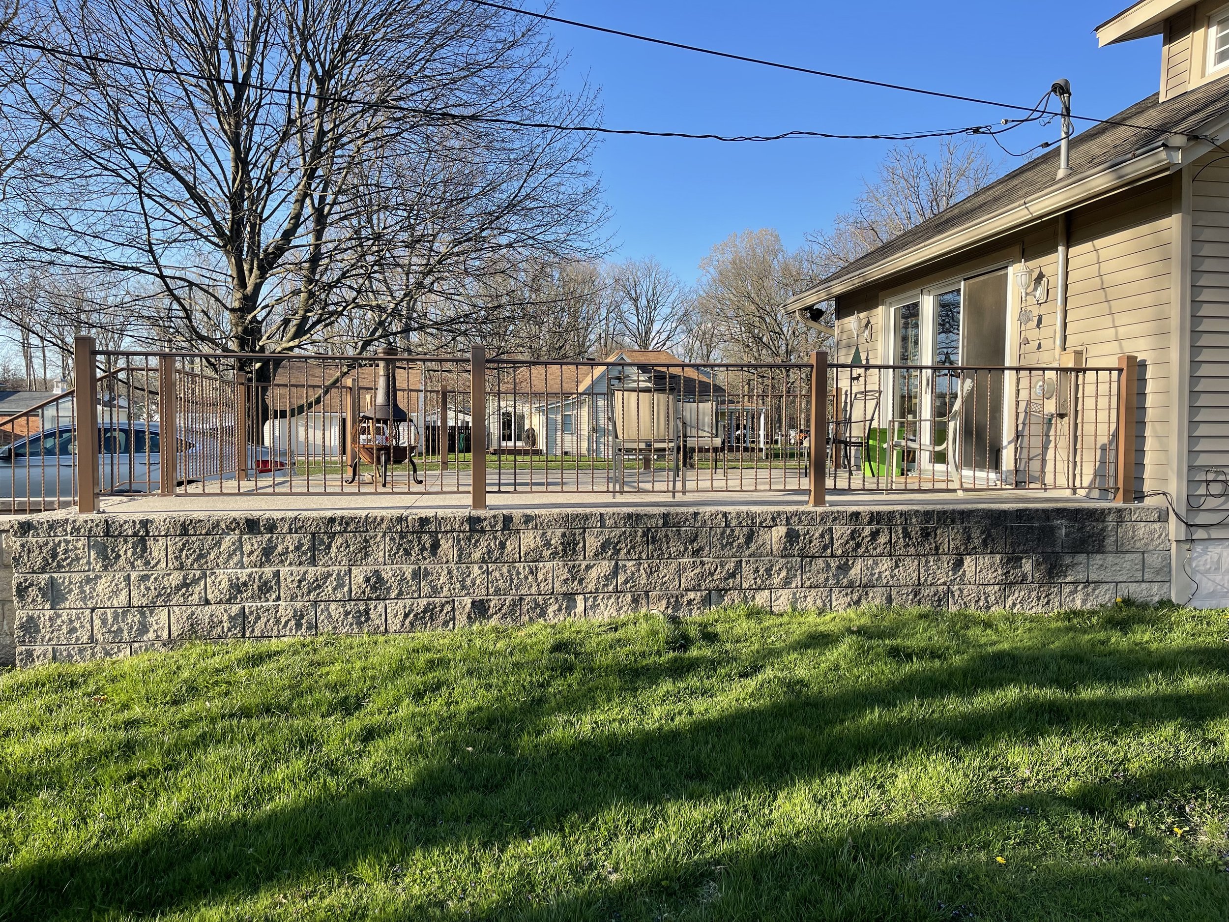 Backyard with a green lawn, a brick wall, a fenced patio with outdoor furniture and a grill, neighboring houses, leafless trees, and a clear blue sky.