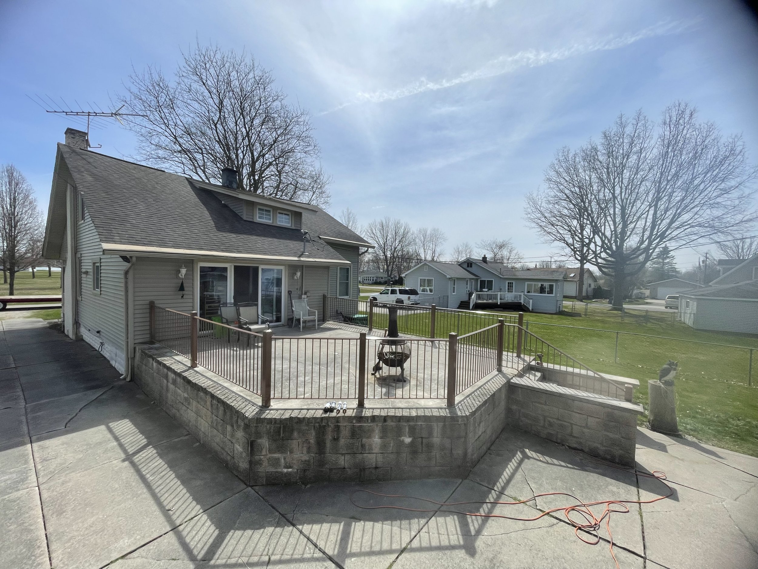 Backyard patio with chairs, a grill, a wooden railing, and a view of neighboring houses and trees under a clear sky.