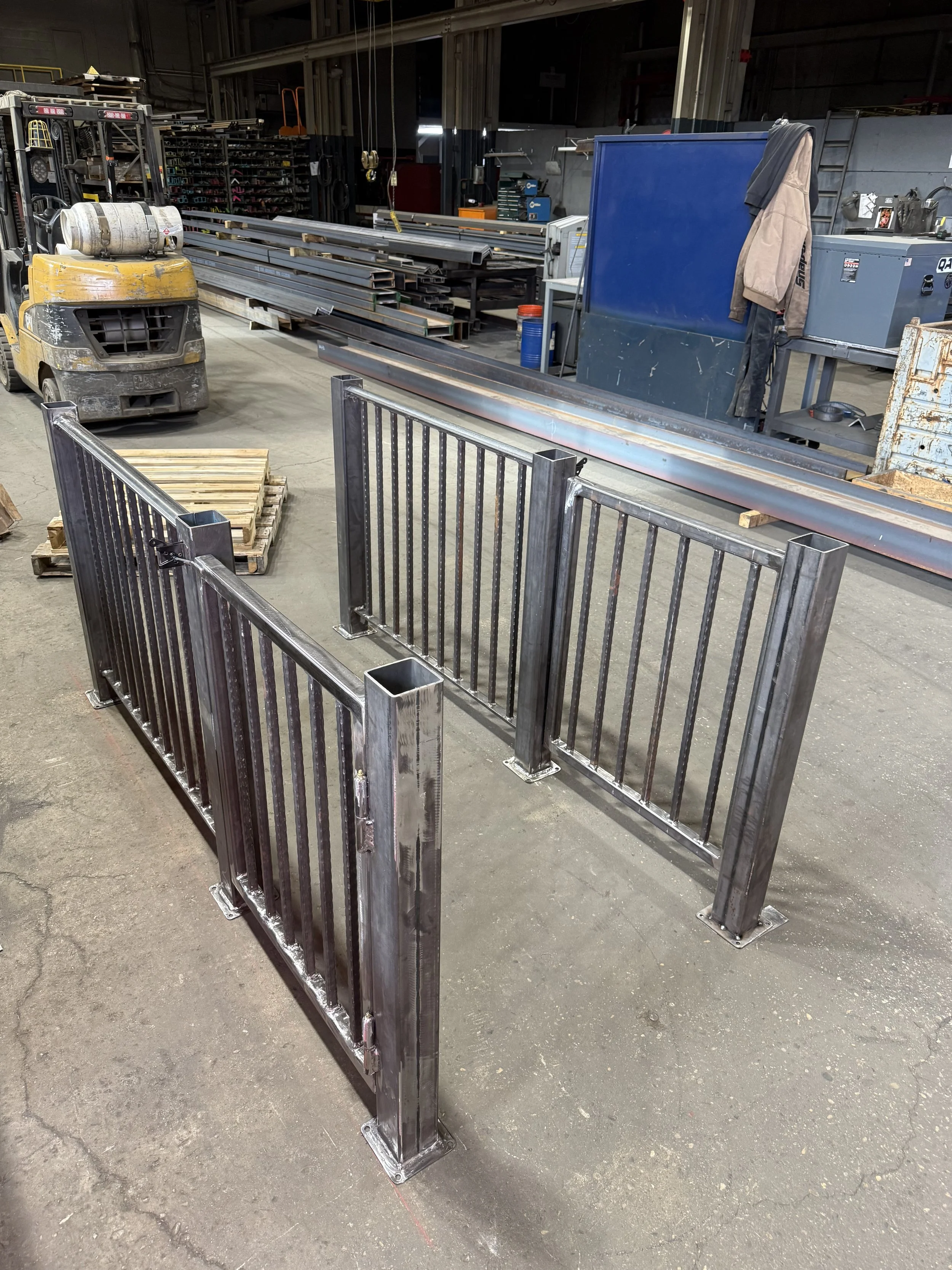 Metal gate in a workshop with metal bars, a forklift, and various tools and materials in the background.