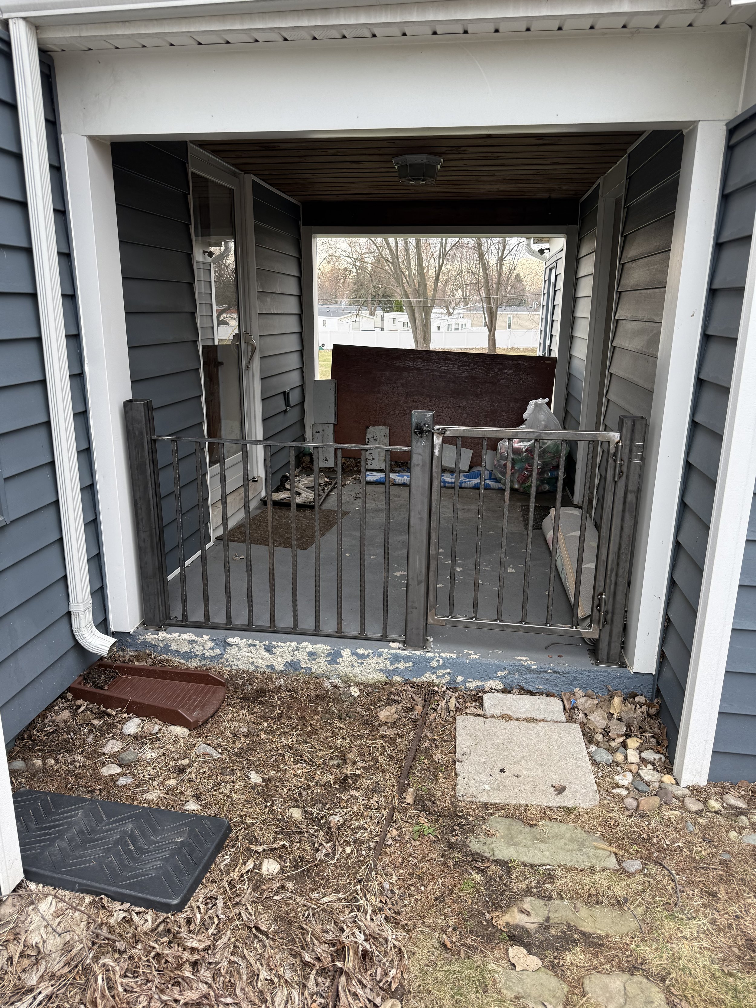 A small covered porch with a metal child gate, a large wooden board, a bag of trash, and various debris. The porch is above ground with a dirt and leaves surrounding it, and the house has blue siding and a white trim. There are stairs leading up to t