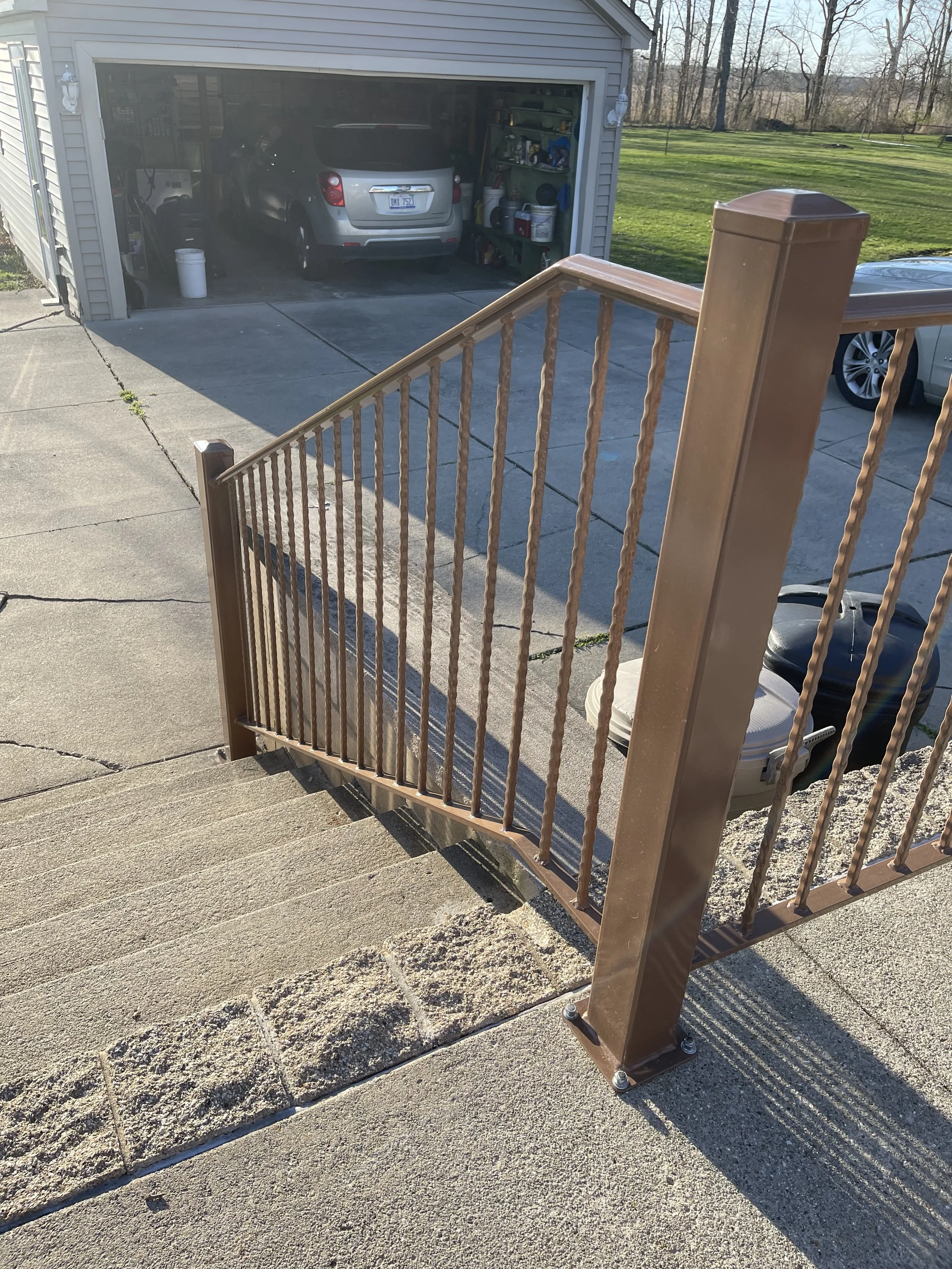 Close-up of a brown metal staircase railing outside, with a concrete patio and a view of a garage with a car inside and gardening supplies, and a grassy yard in the background.