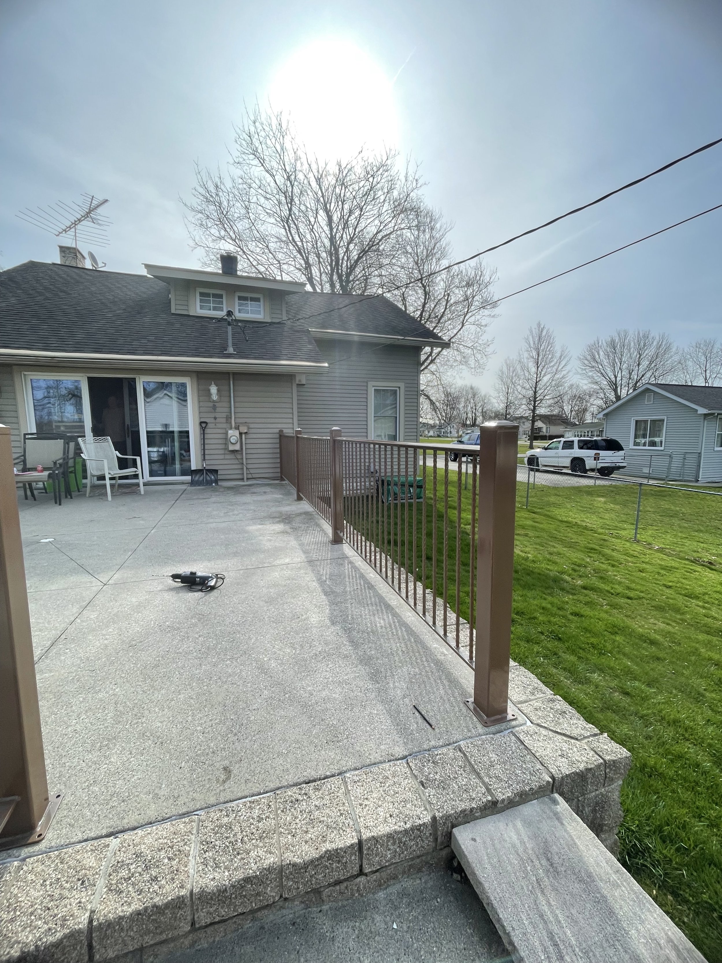 Residential backyard featuring a concrete patio with patio furniture, a wooden and stone step, a grassy area, and neighboring houses with trees in the background under a clear sky with bright sunlight.