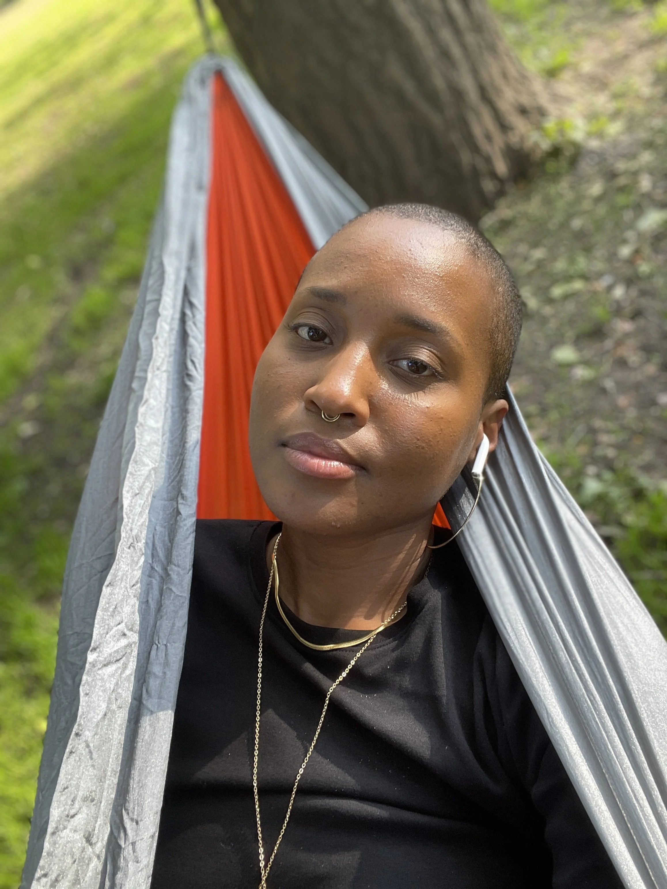 A woman, Janine Bell, with a shaved head laying in a hammock outdoors next to a tree, wearing a black shirt and gold jewelry.