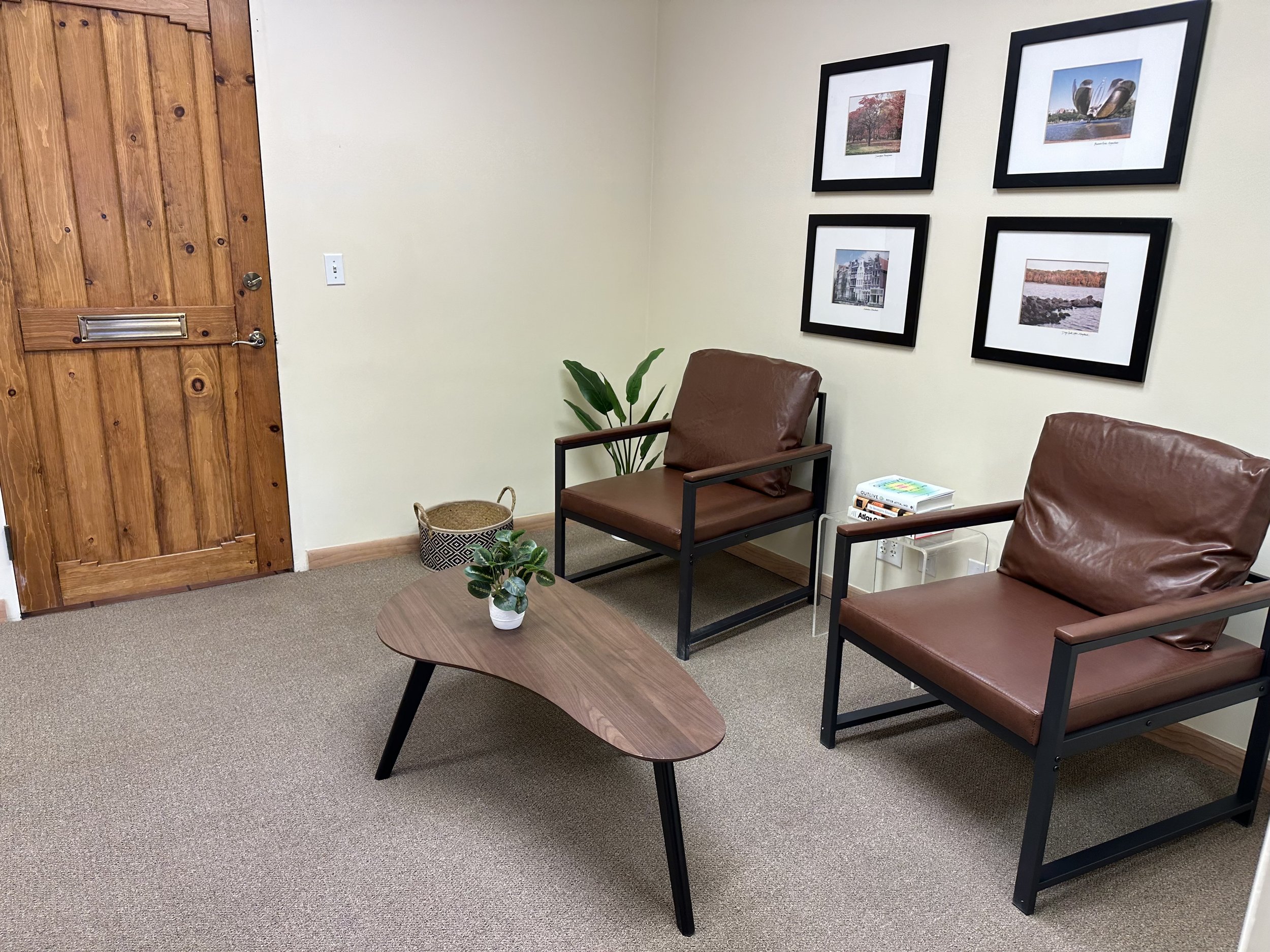 A waiting area with two brown leather chairs with black metal frames, a small wooden coffee table with a potted plant, a basket, and some books, against a wall decorated with four framed landscape photographs and a wooden door.