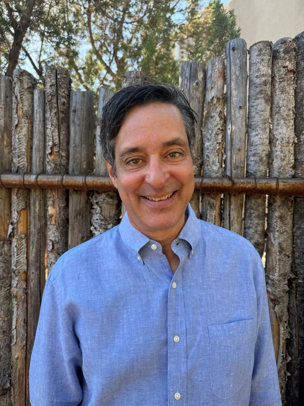 A man with dark hair and a blue button-up shirt smiling in front of a wooden fence with trees in the background.