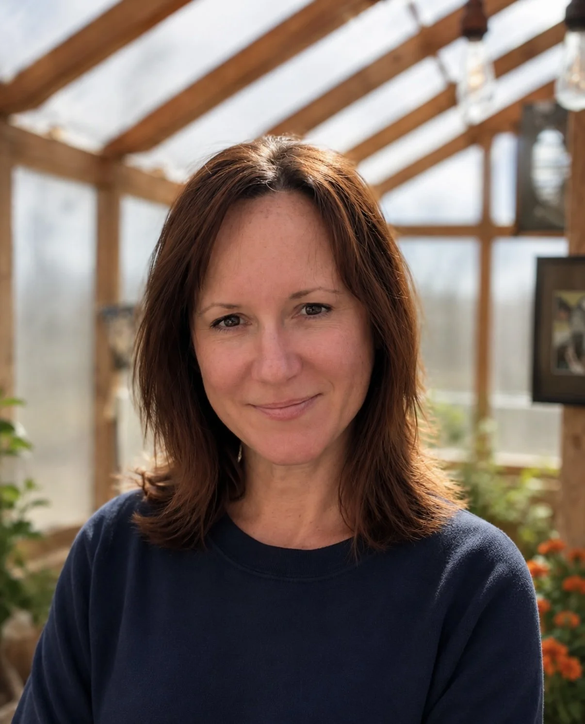 A woman with shoulder-length reddish-brown hair, smiling softly, in a sunlit greenhouse with wooden beams and photos on the walls.