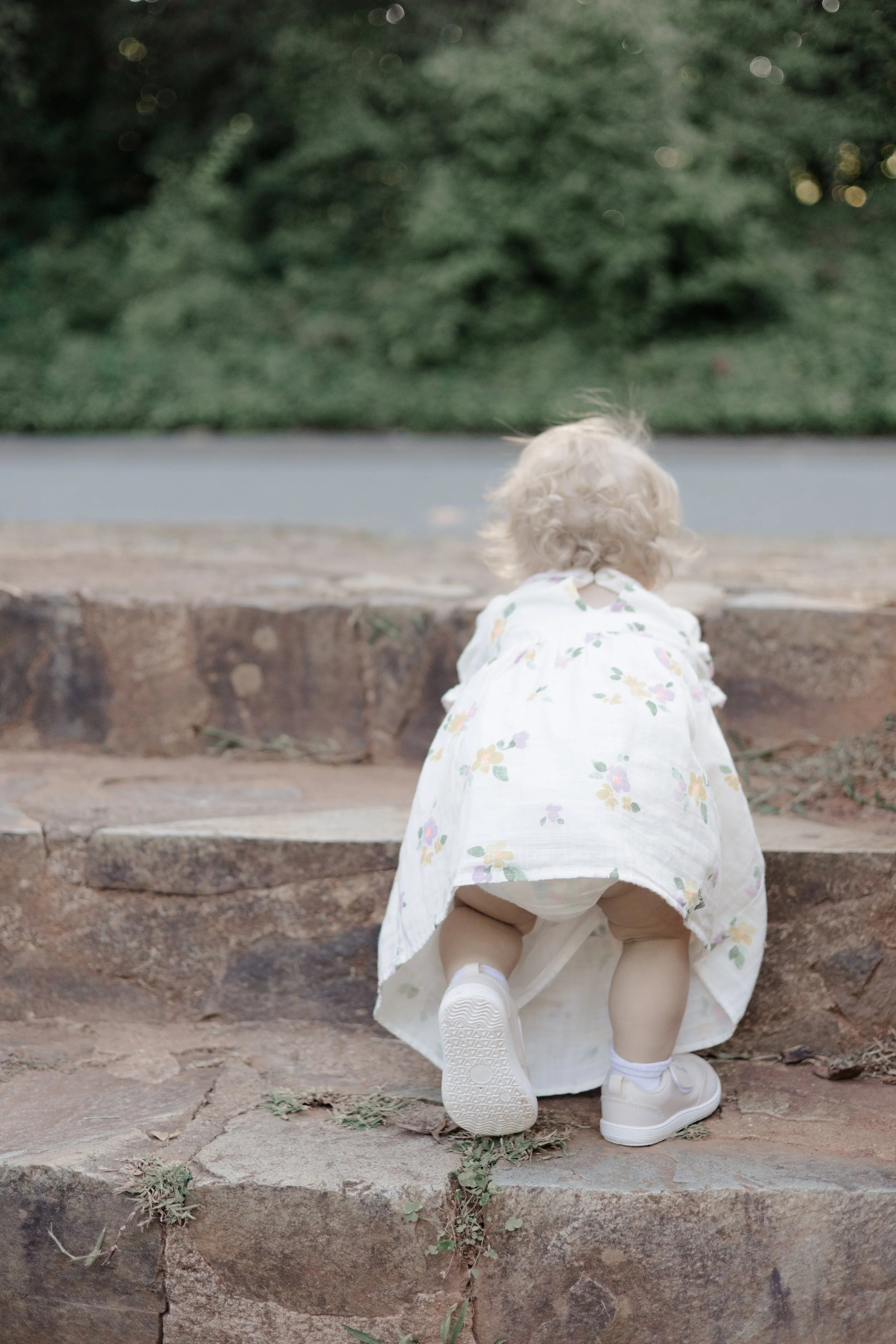 A young child with curly blonde hair crouching on stone steps outdoors, wearing a white floral dress, white sneakers, and white socks, with her back to the camera and greenery in the background.