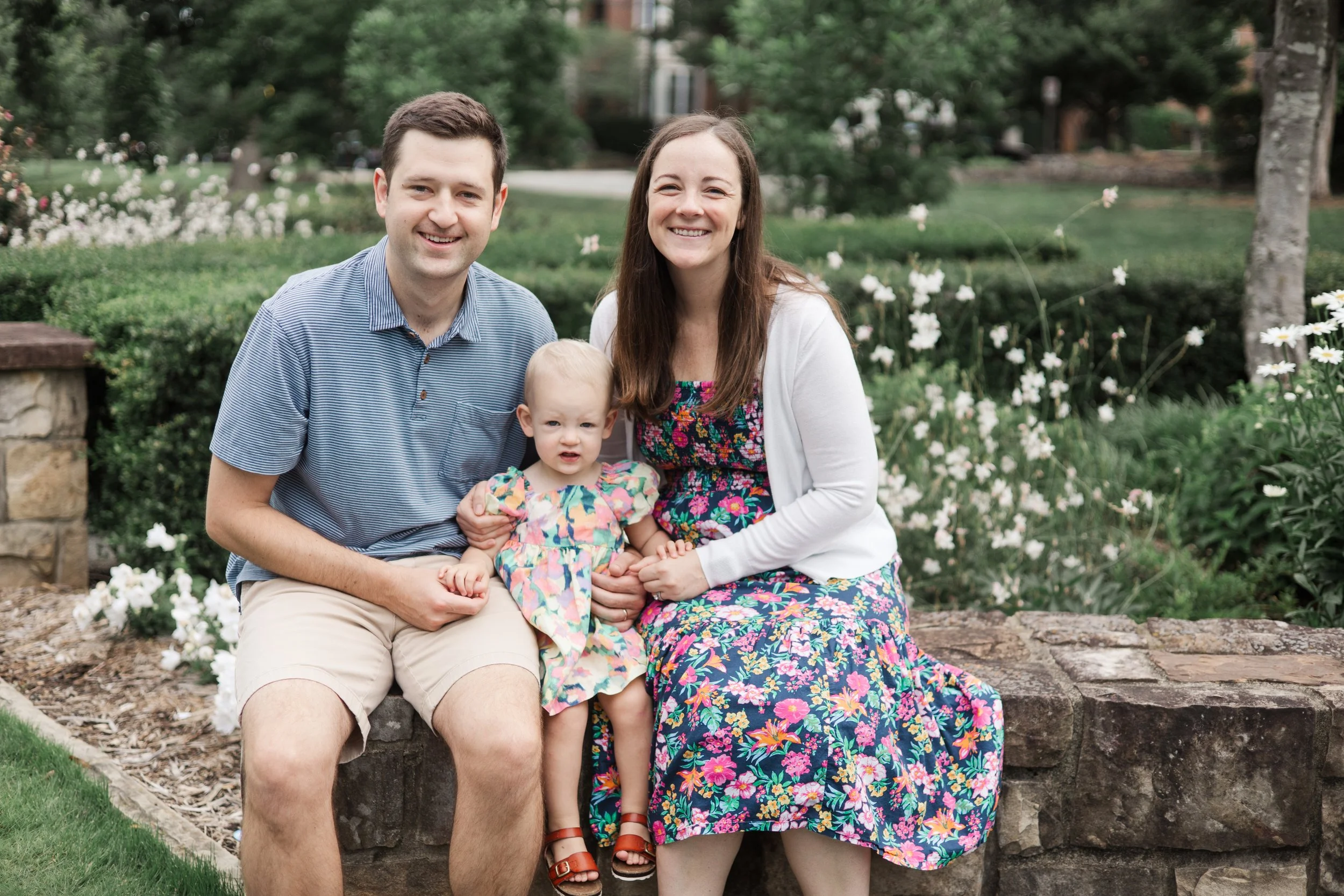 A family of three sitting on a stone bench outdoors in a garden with trees and white flowers, smiling at the camera. The man is wearing a blue striped shirt and beige shorts, the woman is in a colorful floral dress with a white cardigan, and the child is dressed in a brightly colored floral dress.