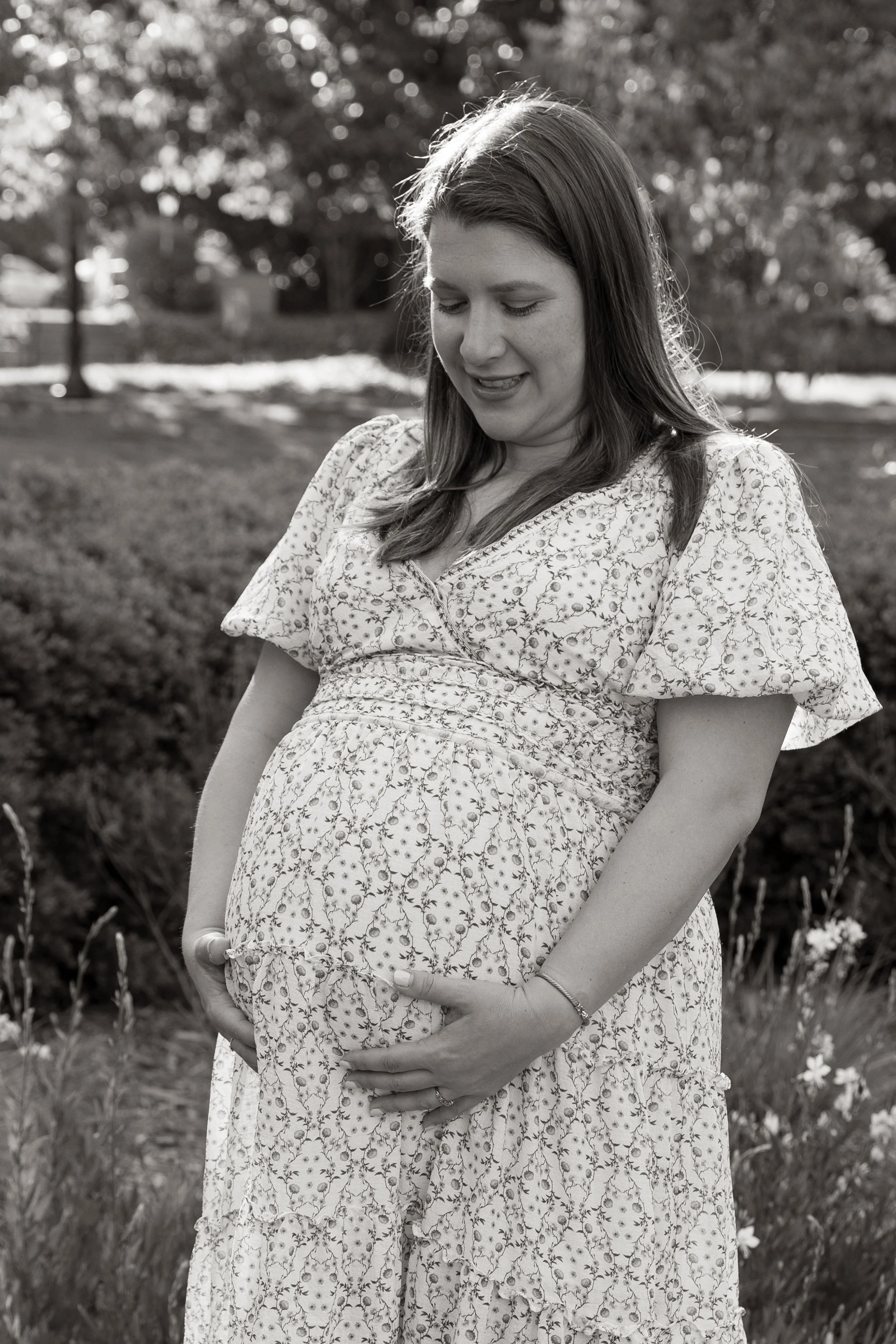 A pregnant woman standing outdoors in a floral dress, gently holding her belly, in a garden setting with trees and flowers.