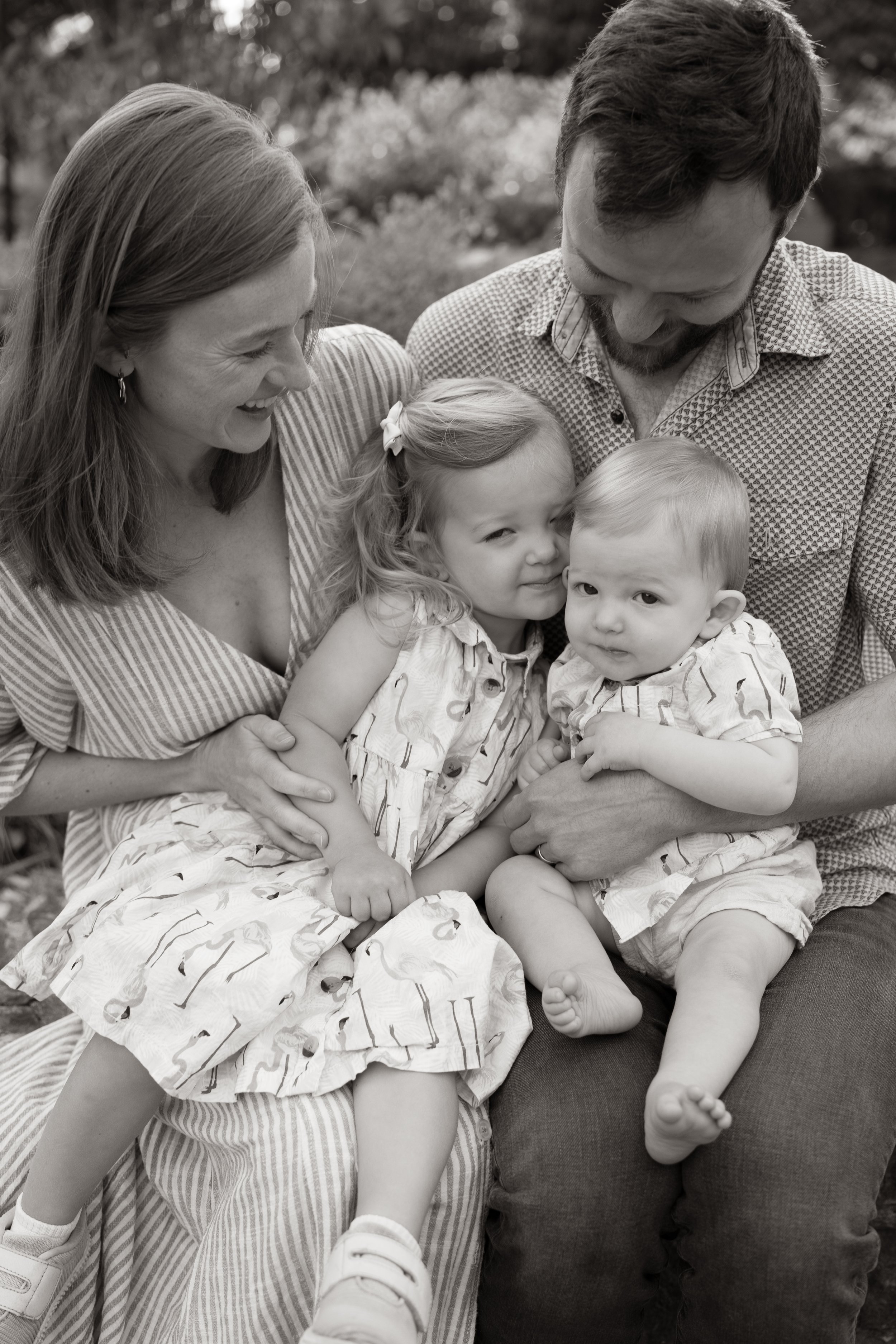 A family of four, including a woman, a man, and two young children, smiling and hugging outdoors on a sunny day.