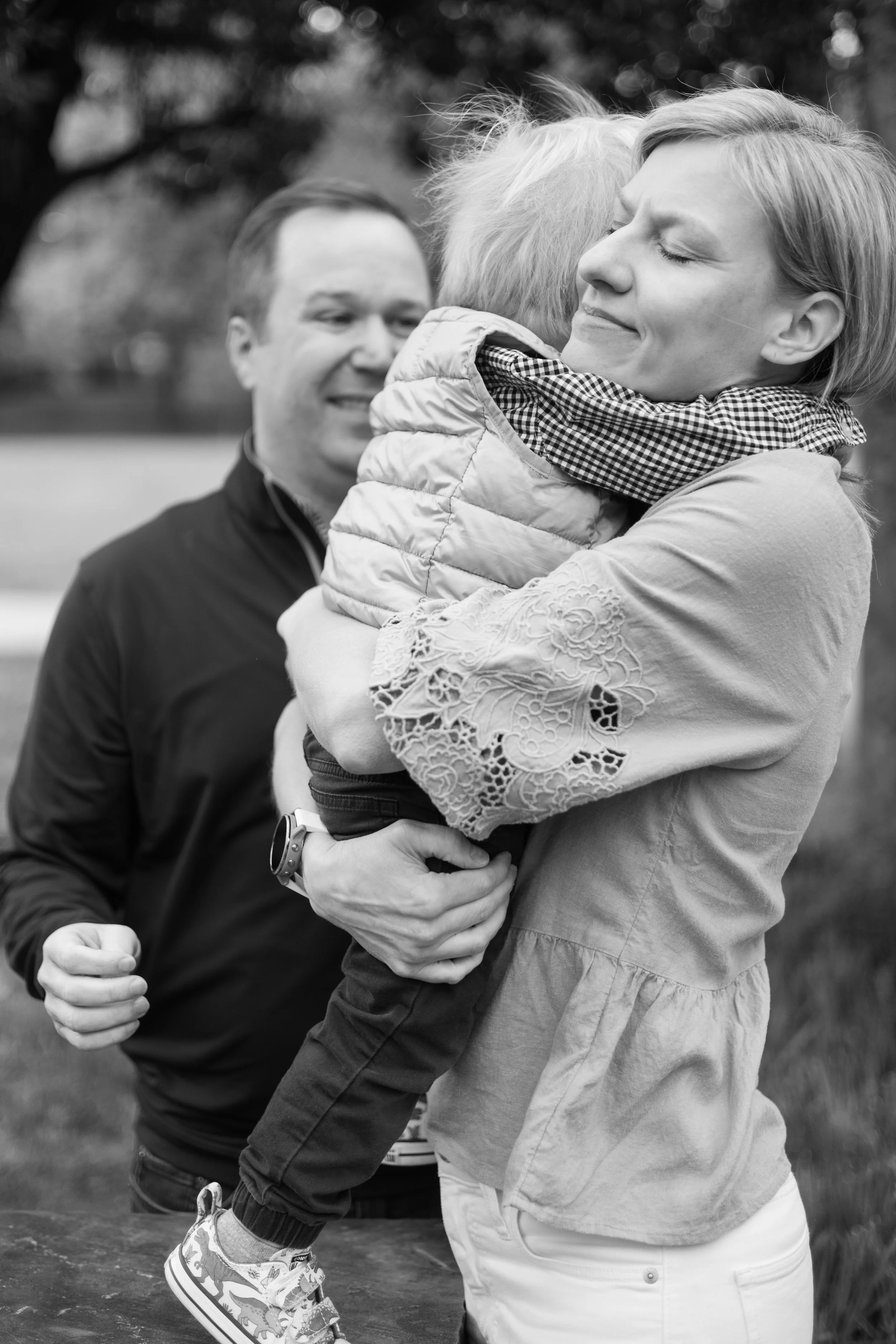 A black and white photo of a woman holding and hugging a young child, with a man in the background. The woman has a slight smile, the child is hugging her tightly, and the man appears to be smiling or laughing.