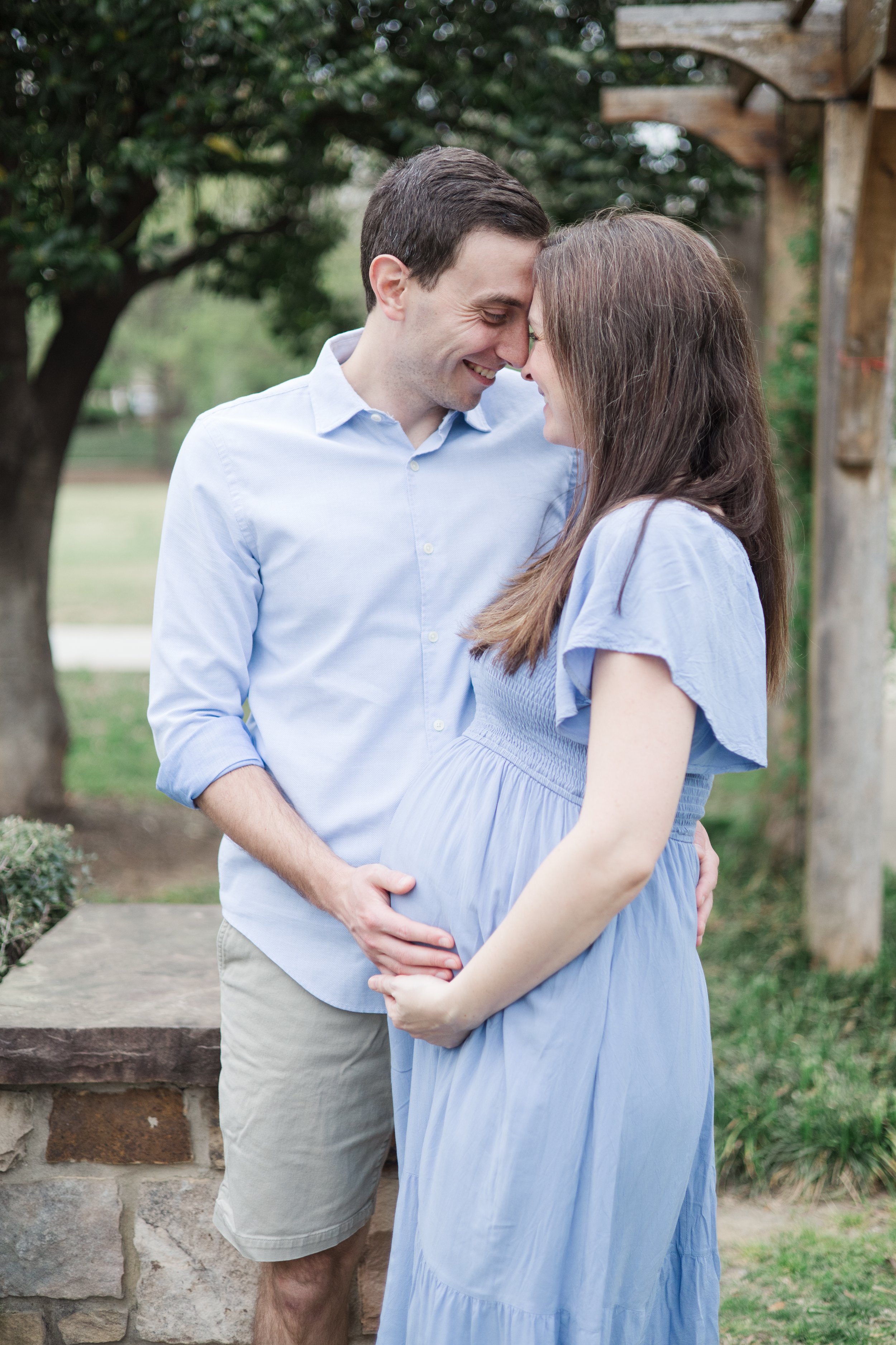 A pregnant woman and a man are standing close together outdoors, with their foreheads touching and eyes closed, smiling, in a park or garden setting.
