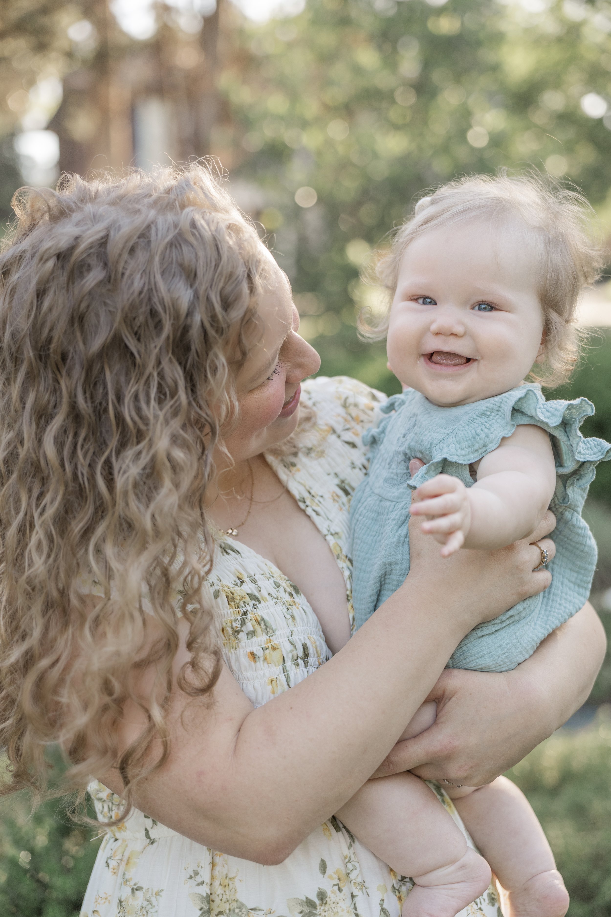 A woman with curly blonde hair holding a smiling baby with curly hair outdoors with blurred trees in the background.