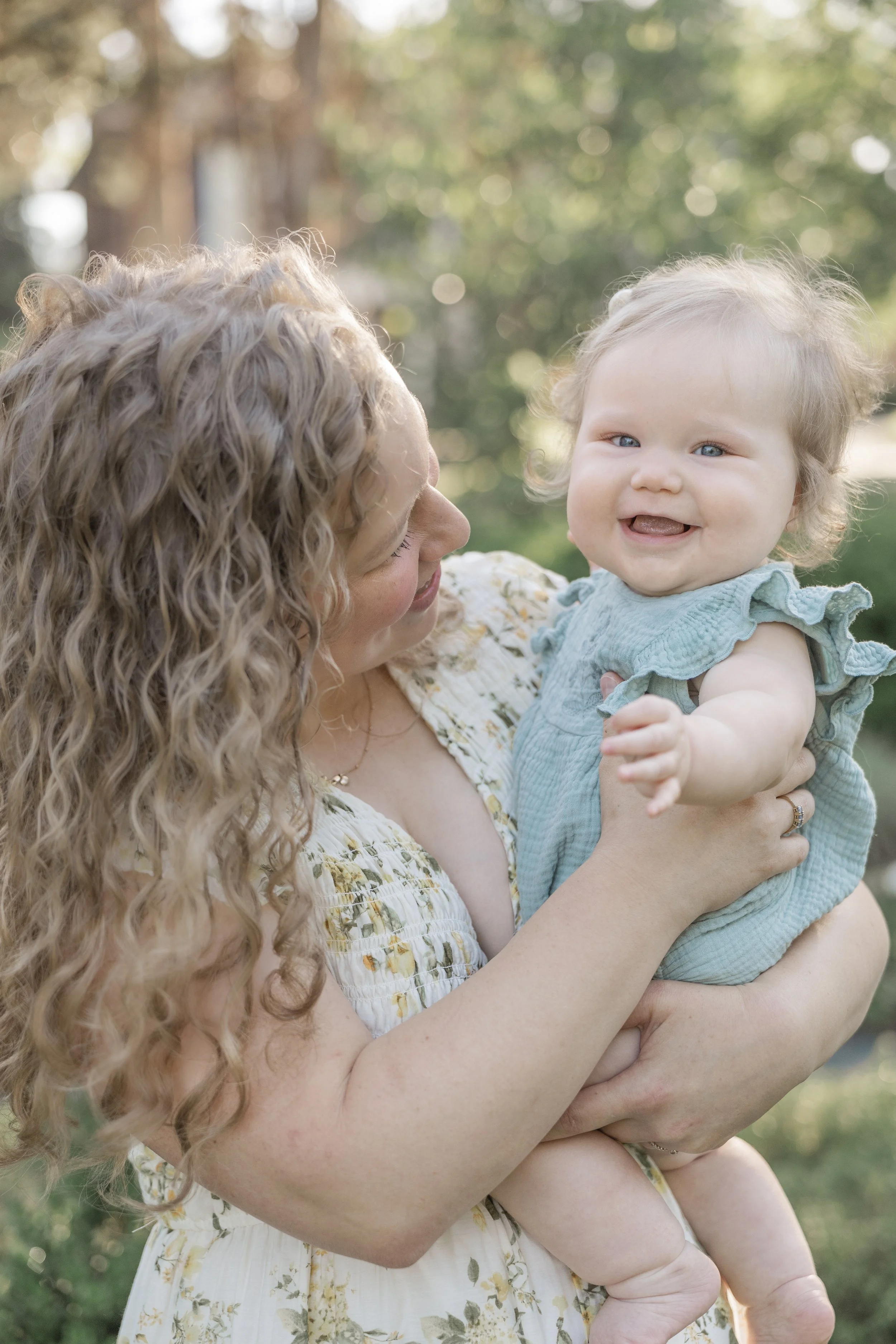 A woman with curly blonde hair holding a smiling baby with curly hair outdoors with a blurred background of trees.