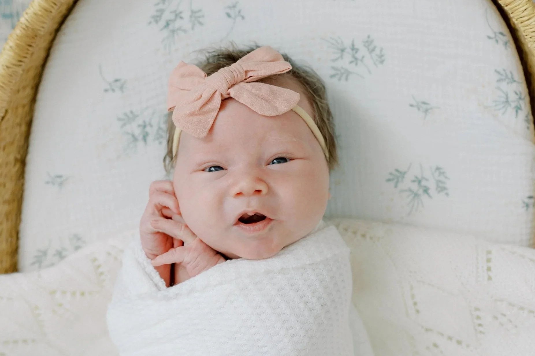 A baby girl lying in a crib with a light pink bow headband, making an expression with her mouth open and eyes slightly squinted.