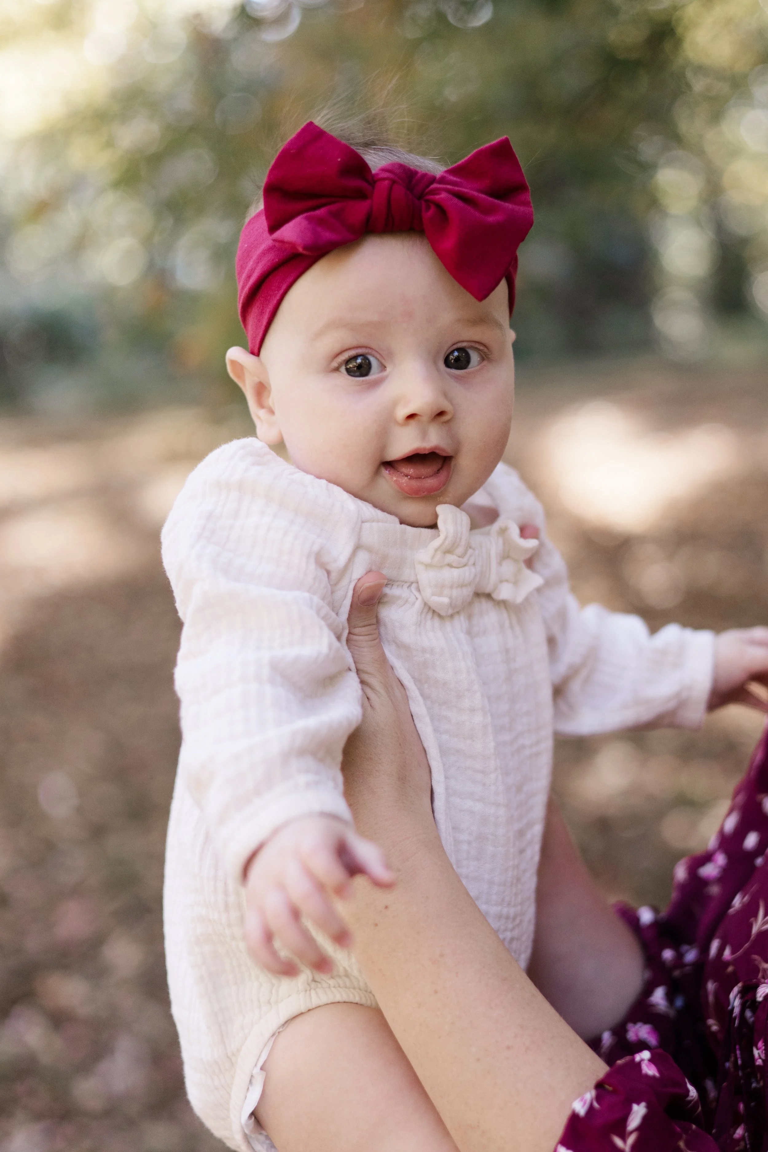 A baby with a big red bow headband, wearing a cream-colored top, being held outdoors, with blurred green and yellow foliage in the background.