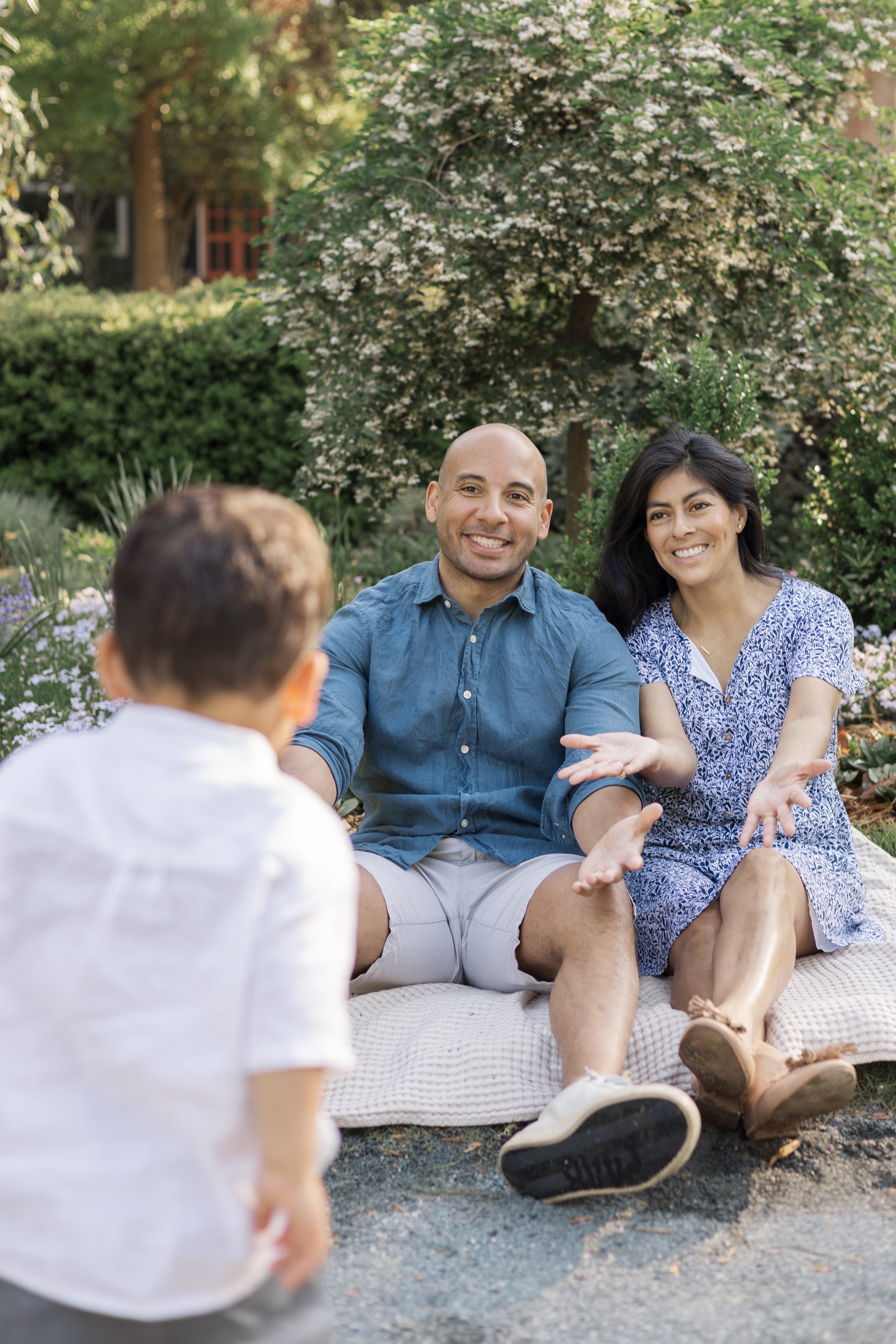 A couple sitting on a blanket in a garden, talking to a young child whose back is visible, surrounded by greenery and flowering bushes.