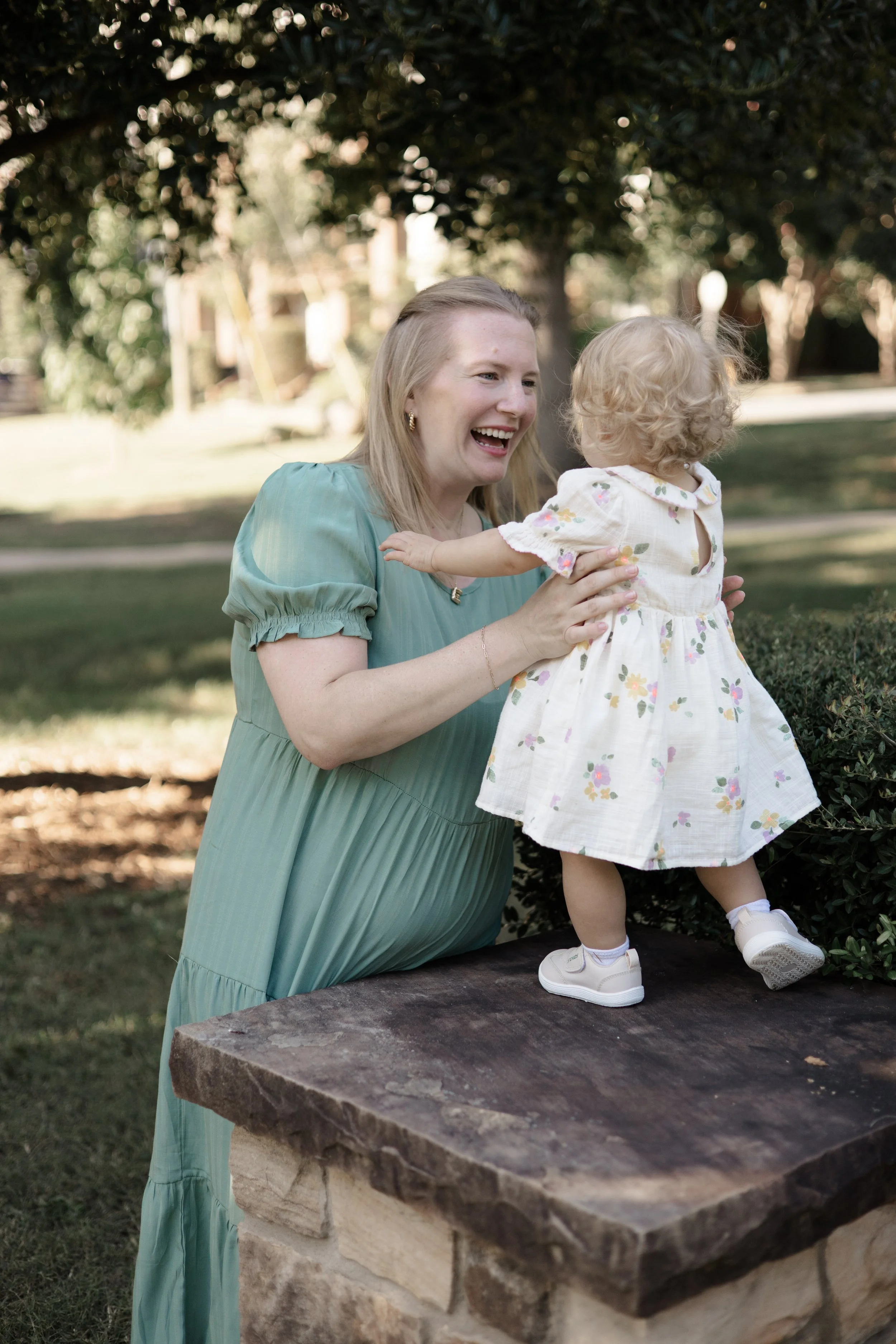 A woman smiling and holding a young girl, who is standing on a stone table outdoors near a tree with a blurred background of greenery and a park.