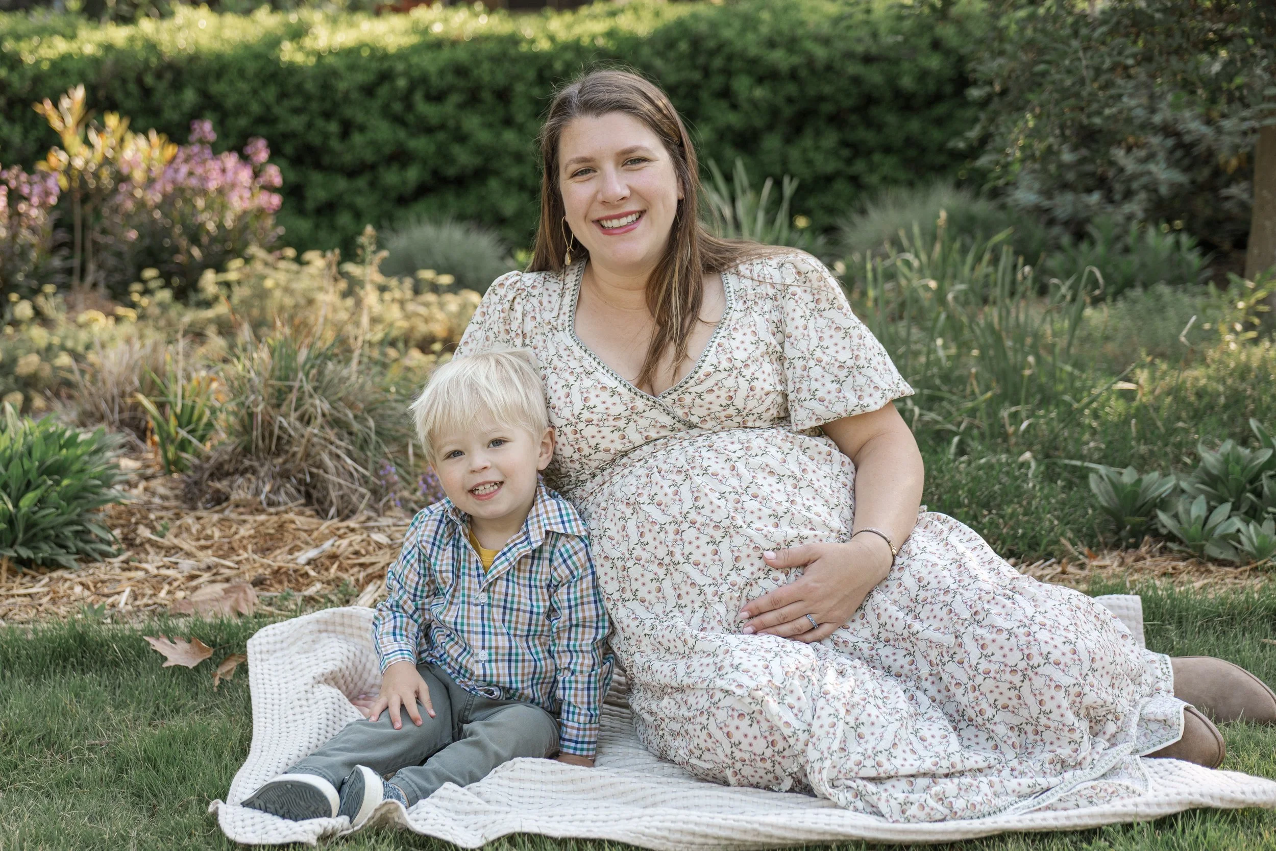 A woman in a floral dress sitting on a blanket outdoors with a young boy, surrounded by green bushes and plants in a garden.