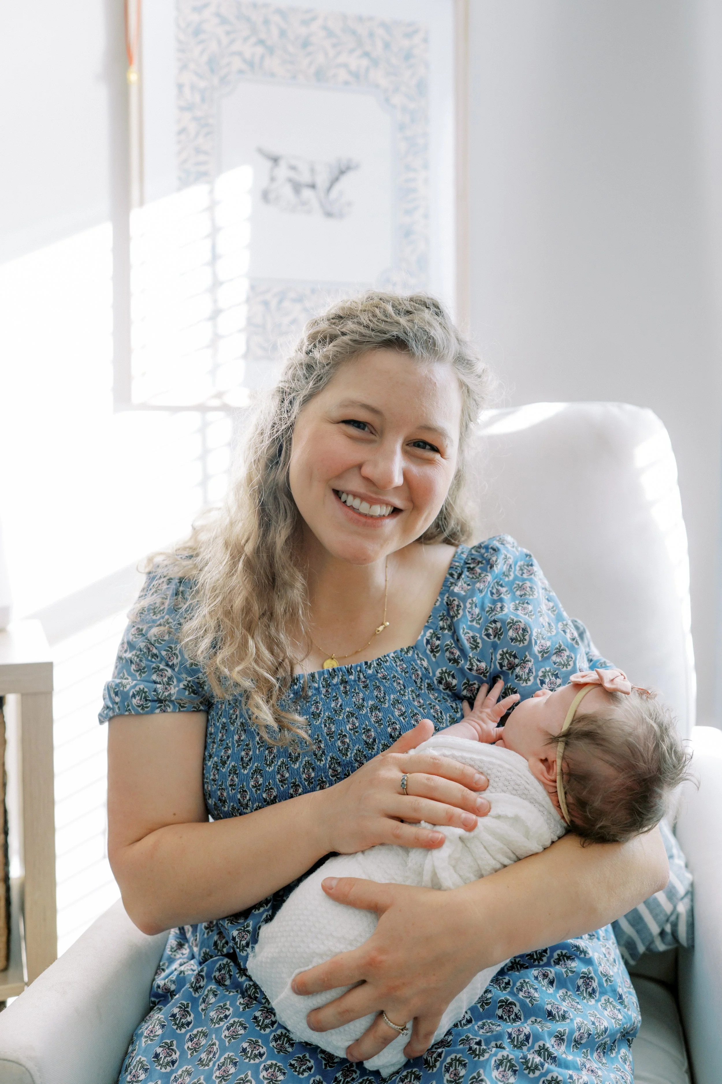 A woman smiling and holding a newborn baby in a bright room with a window and artwork in the background.