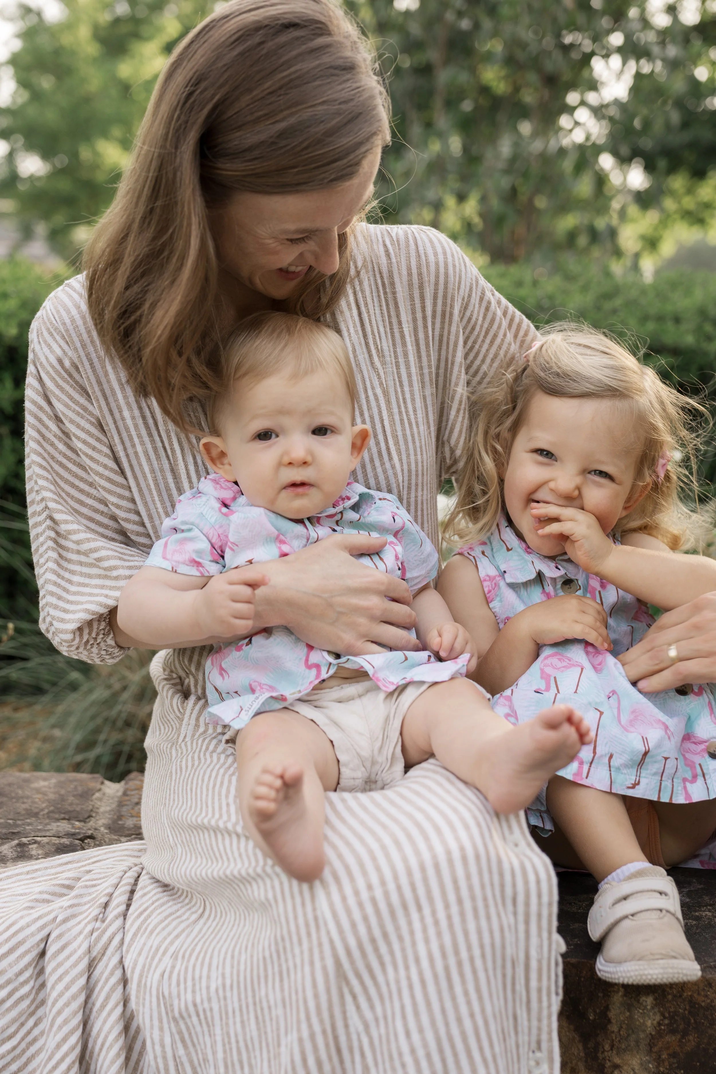 A woman with brown hair smiling and looking down at two young children sitting on her lap outdoors with green trees in the background. The children, a boy and a girl, are wearing matching pastel-colored outfits, and the girl is playfully sticking her tongue out.
