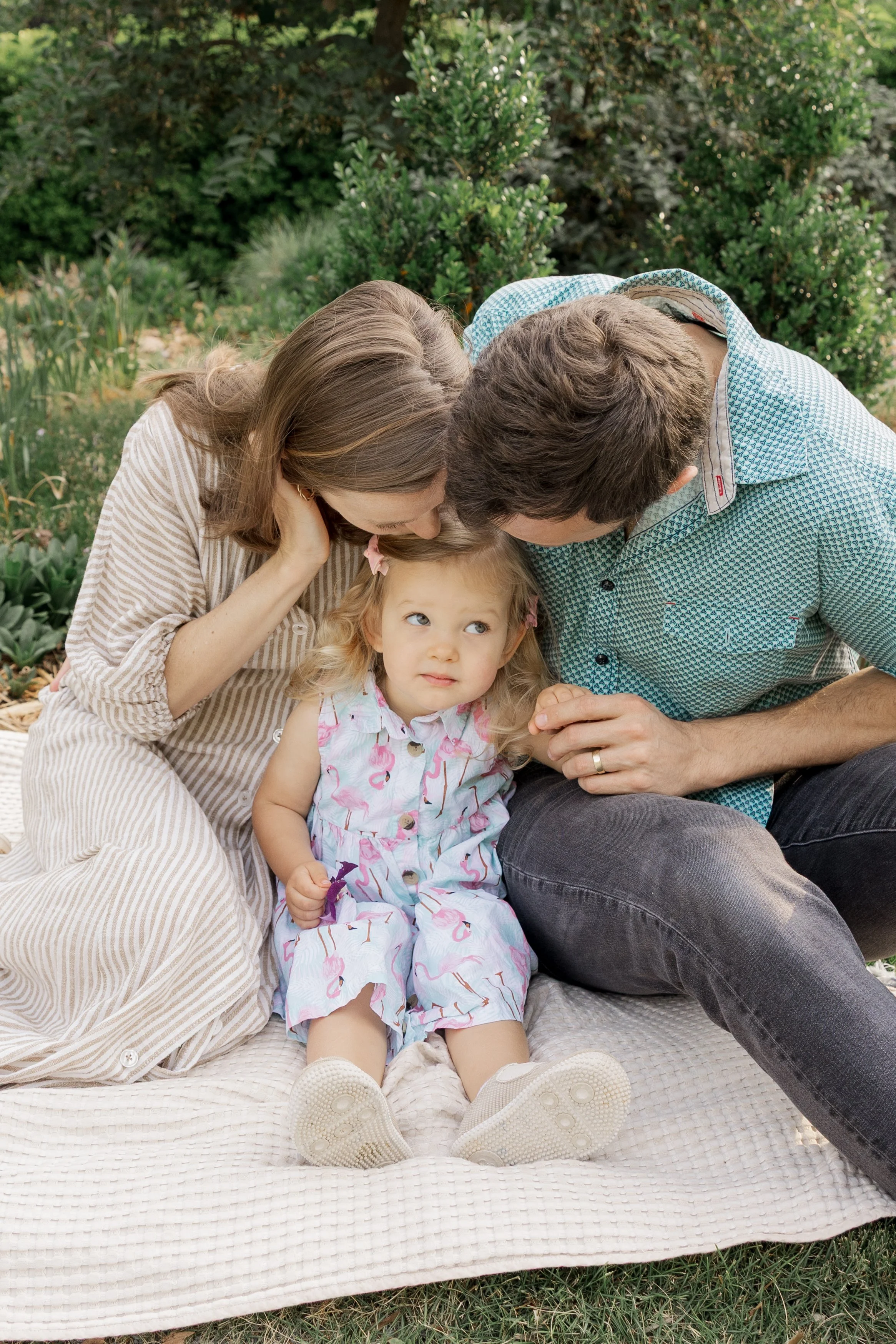 A family of three sitting on a blanket outdoors, with parents kissing their young daughter on the head. The girl has curly blonde hair, is wearing a colorful dress with flamingo designs, and white sneakers. The background features green bushes and tr
