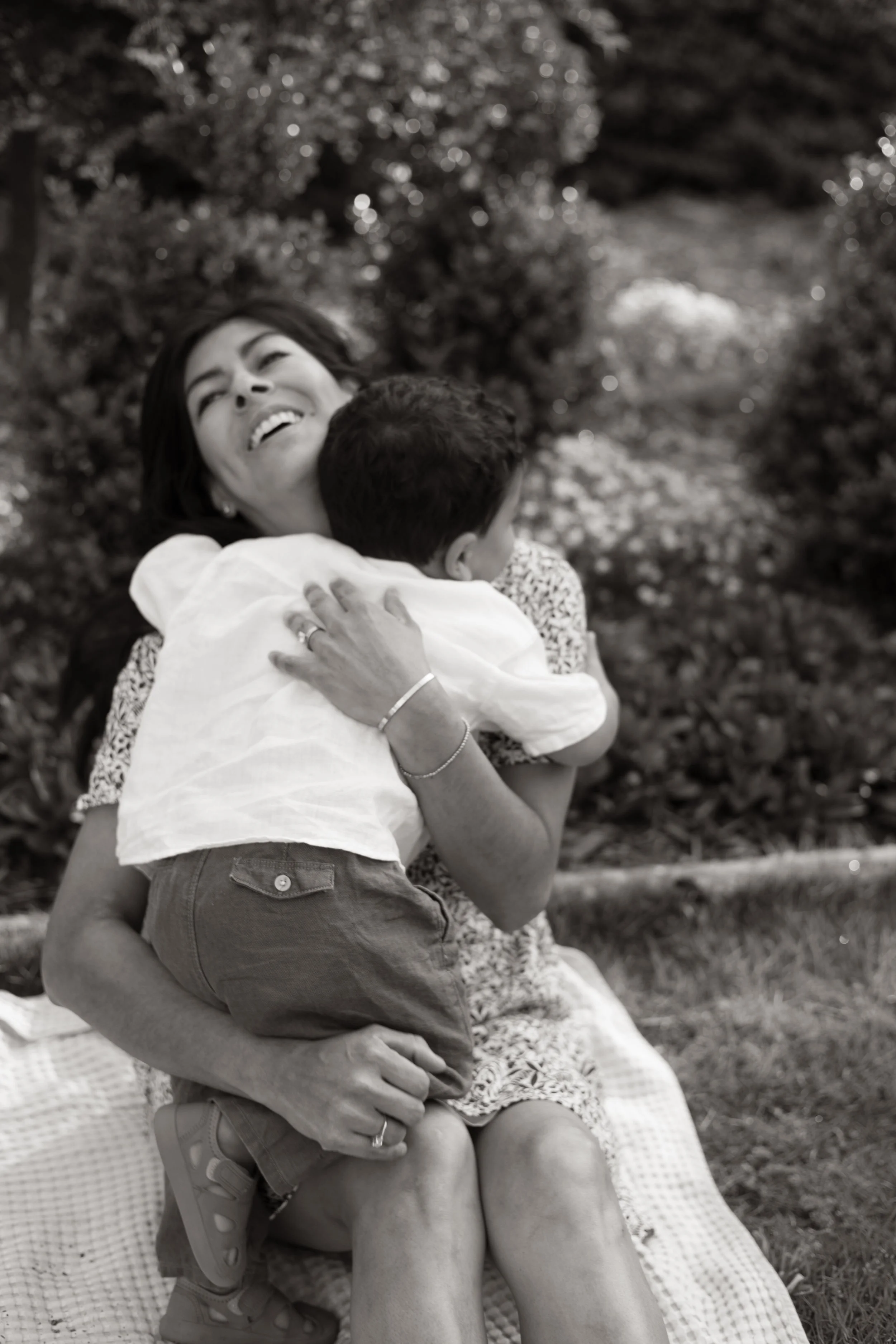 A woman sitting outdoors on a blanket hugging a young boy, both smiling and enjoying a sunny day.