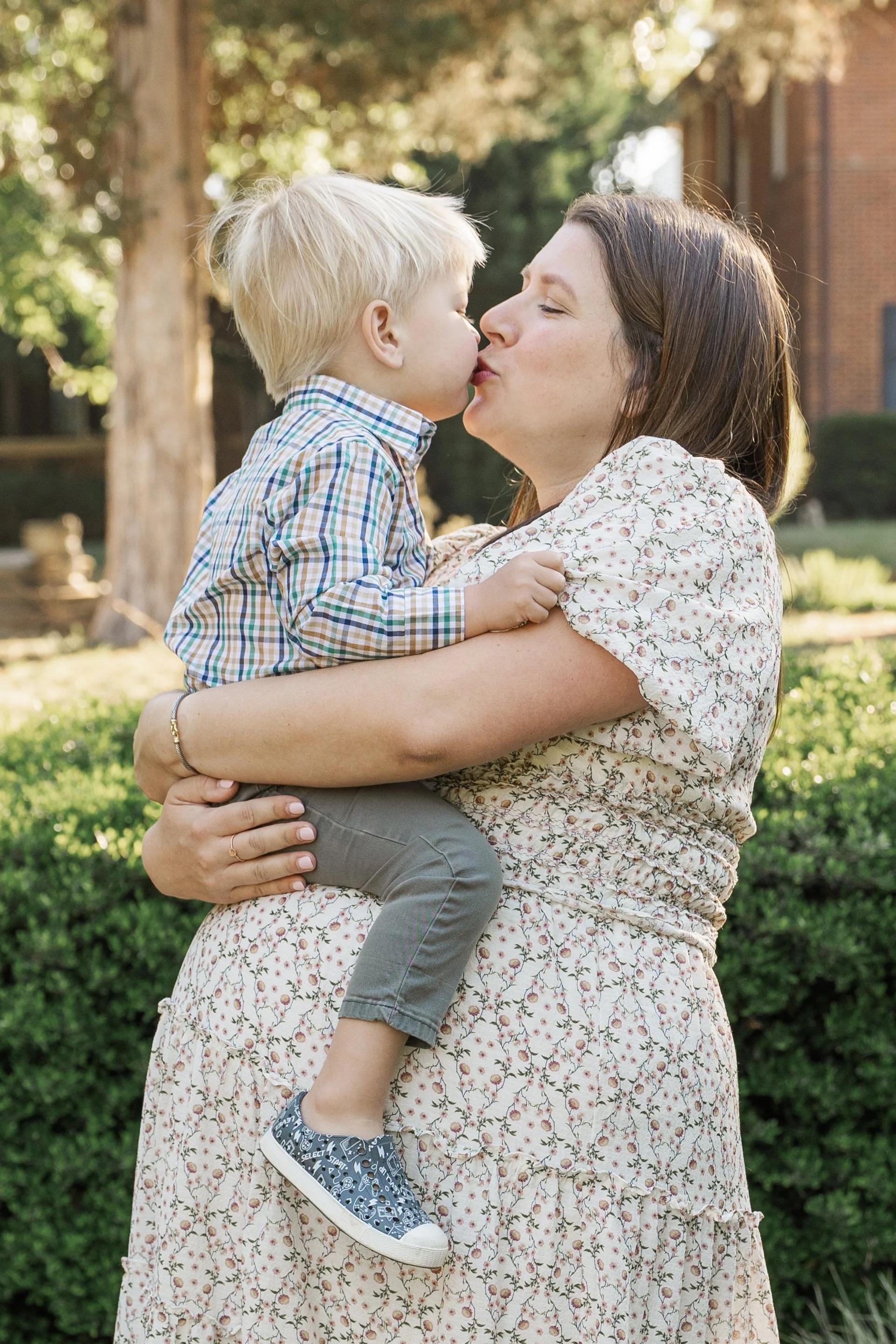 A woman holding a young boy outdoors, about to kiss him, with trees and greenery in the background.