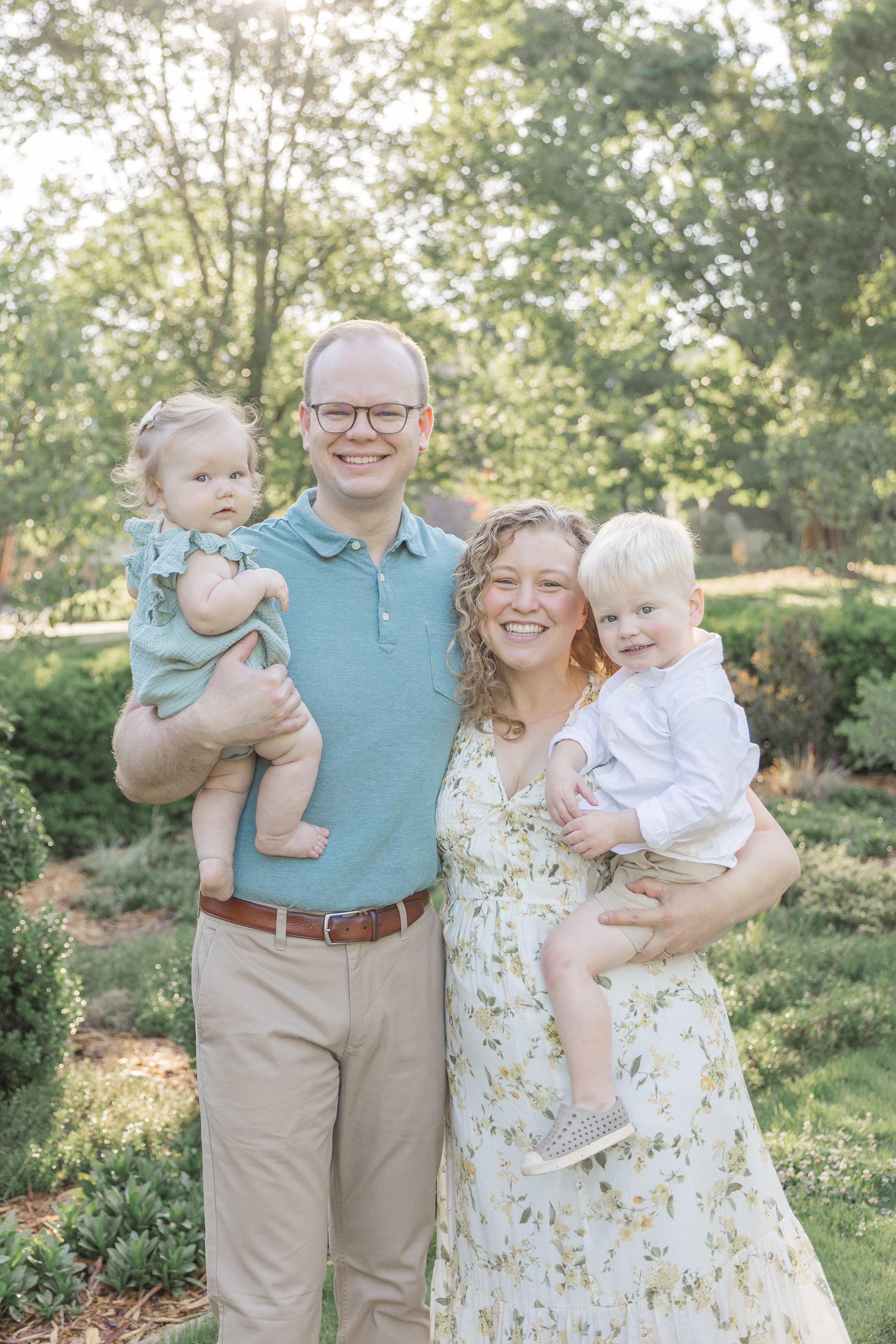 A family of four outdoors in a park with trees in the background. The father is holding a young girl, and the mother is holding a young boy. Everyone is smiling and dressed in light-colored, casual clothing.
