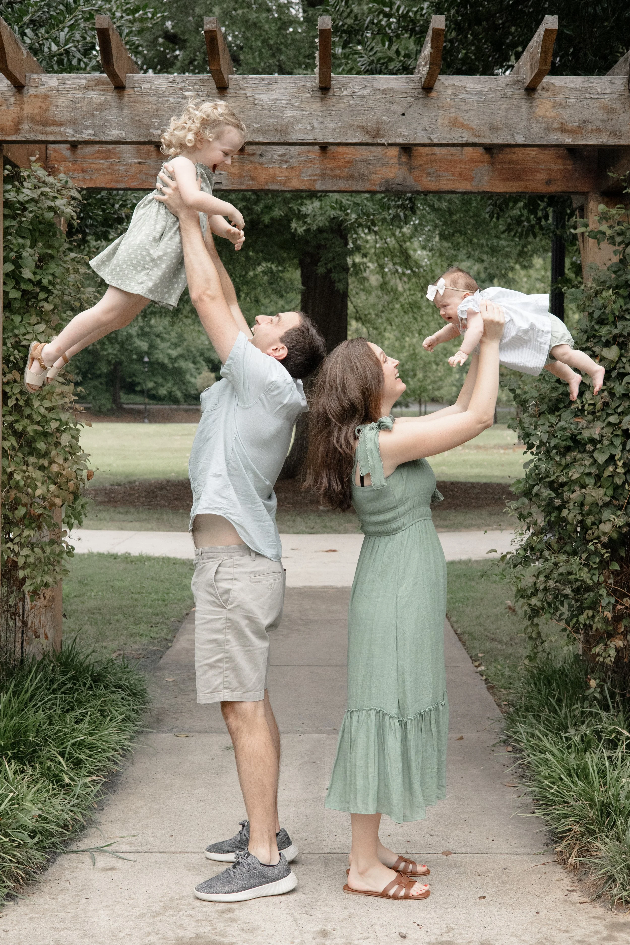 A man and a woman each lifting a young girl and a baby, respectively, in an outdoor park setting with greenery. The man is smiling and supporting the older girl, who is reaching down towards the baby. The woman is smiling and holding the baby, who appears to be laughing.