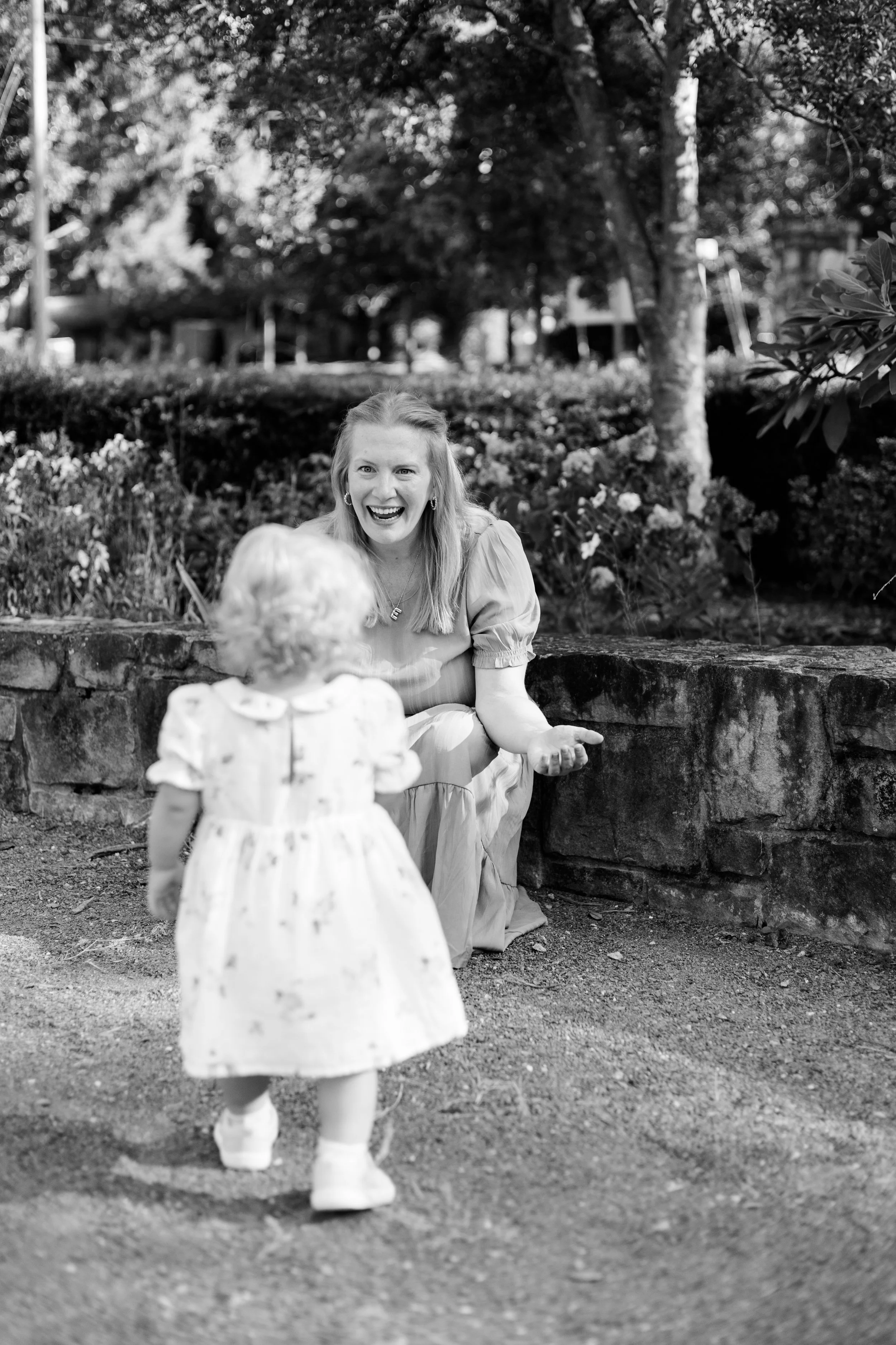 A woman with long hair and earrings, smiling and sitting by a stone wall in a park with trees and flowers in the background, as a small girl in a dress approaches from in front of her.