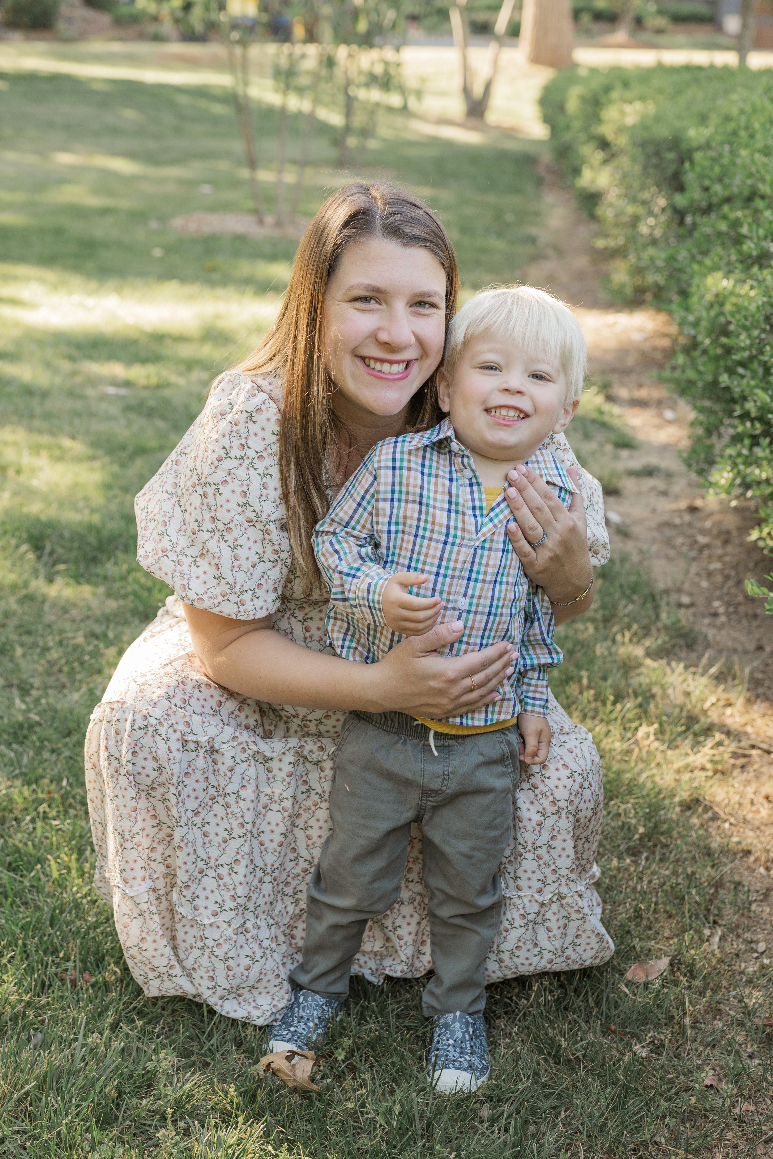 A woman and a young boy smiling and hugging outdoors in a park or garden with green grass and trees in the background.