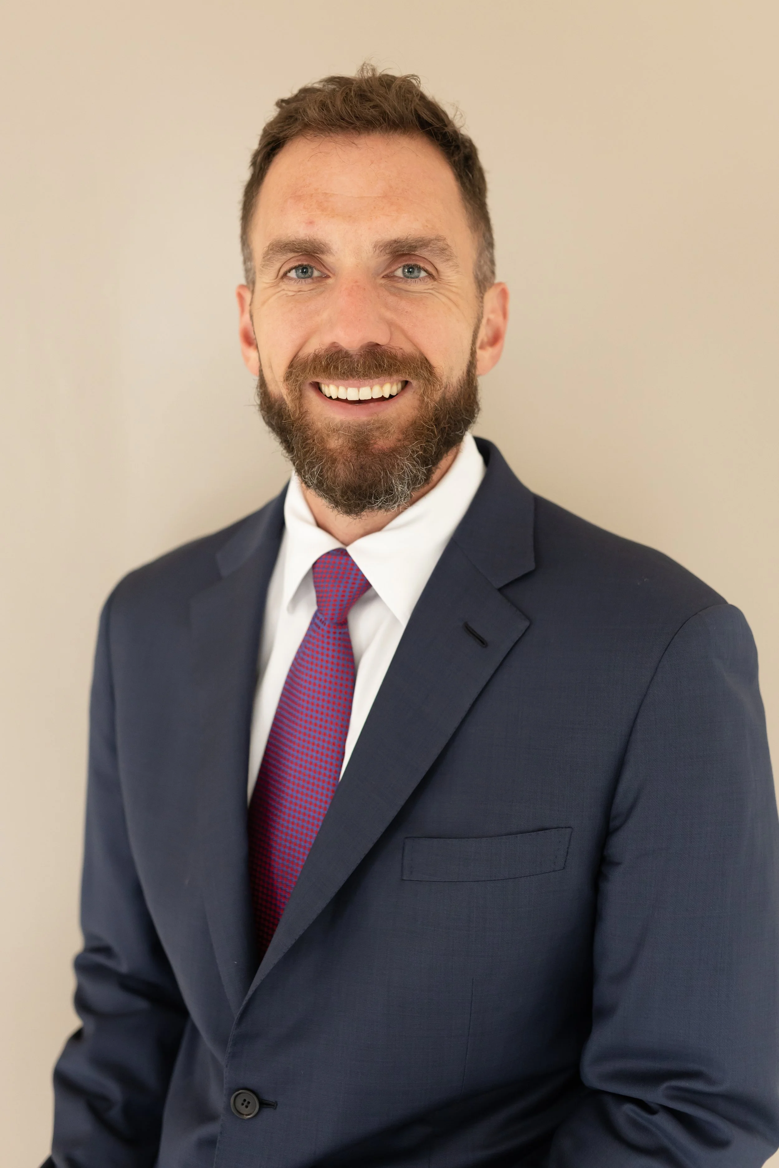 A smiling man with a beard in a navy suit, white shirt, and red tie, standing against a plain light-colored background.