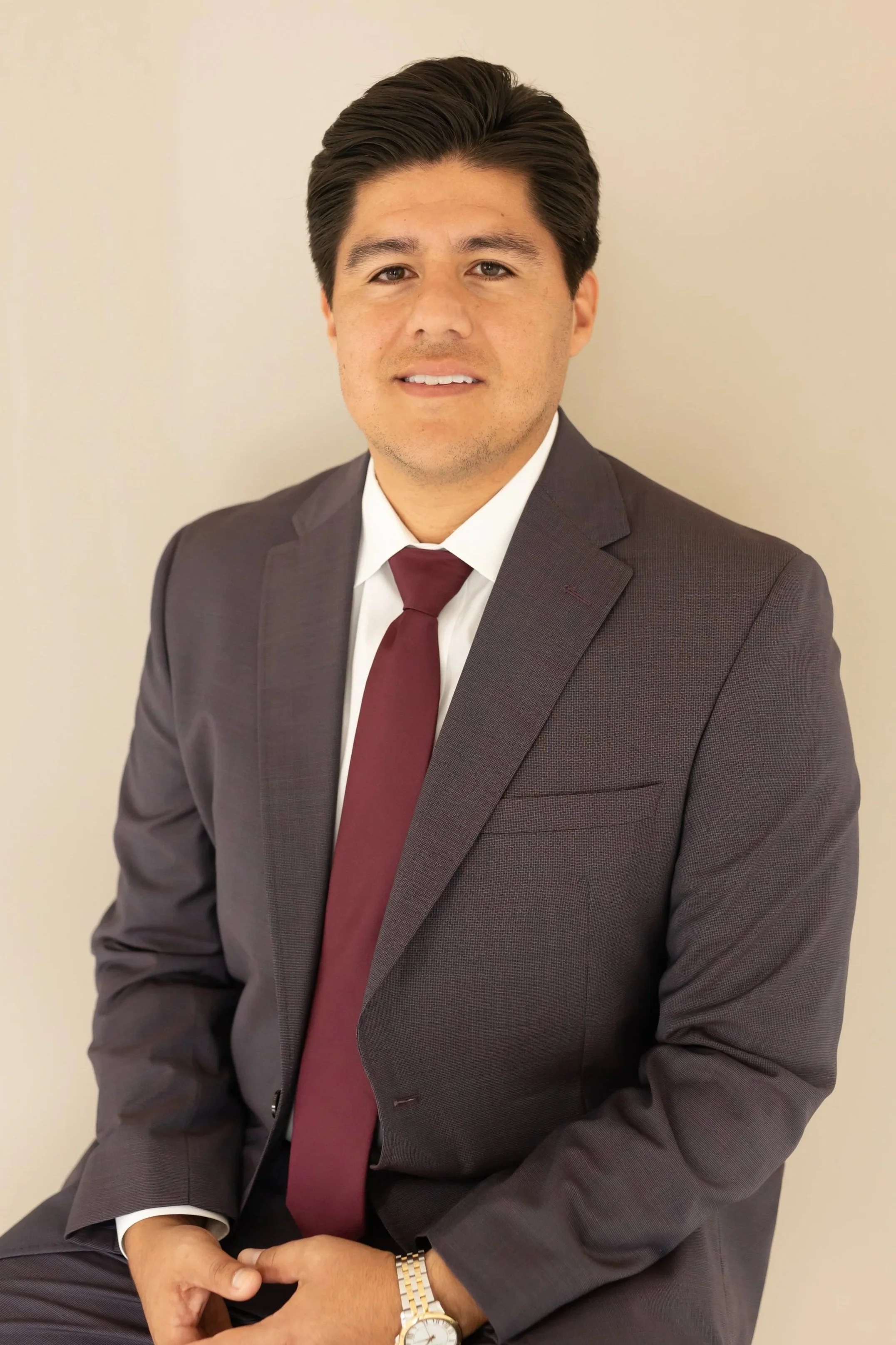 A man wearing a dark gray suit, white shirt, and maroon tie, sitting against a plain light-colored wall.