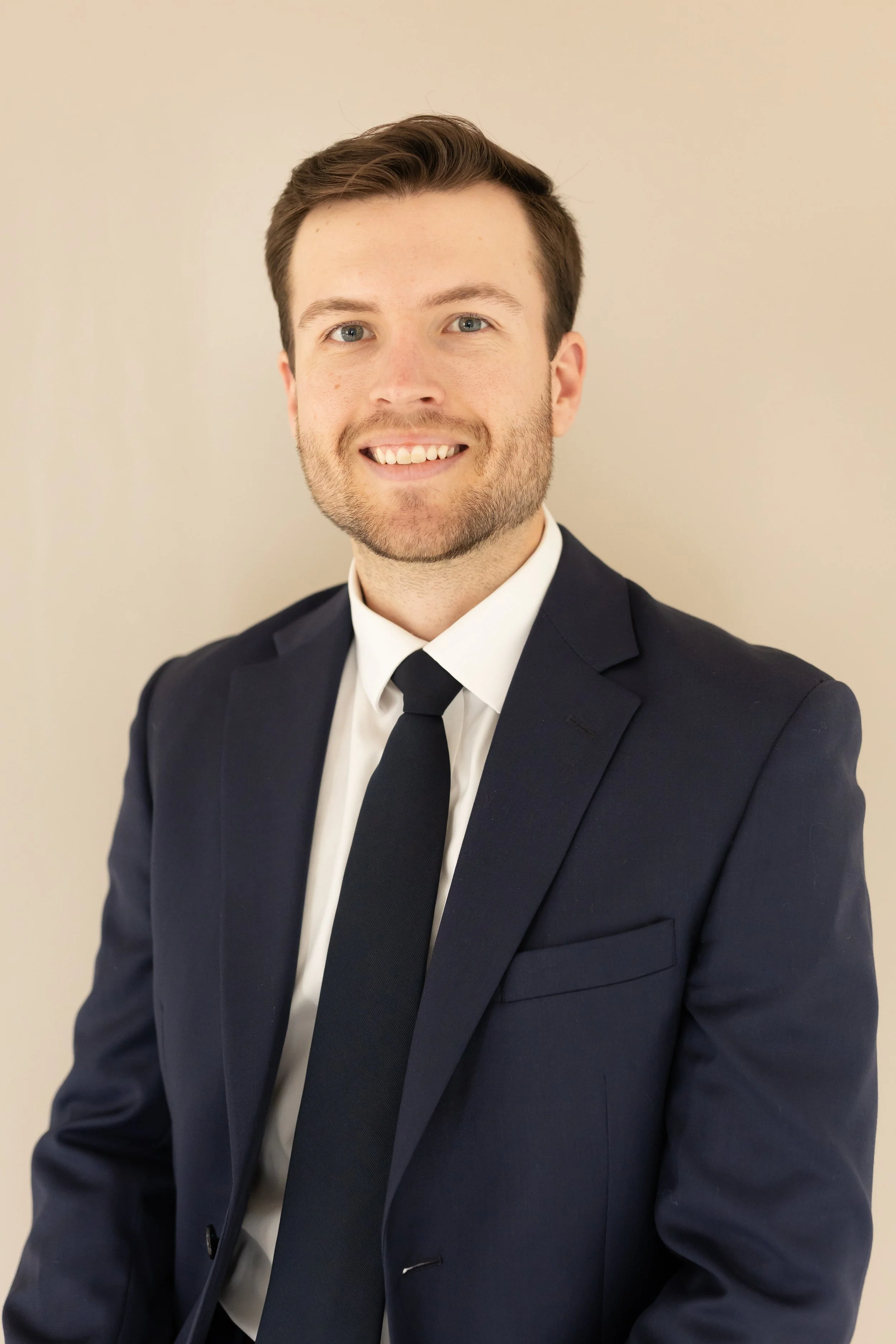 A young man with brown hair and blue eyes, smiling, wearing a navy suit, white shirt, and dark tie, standing against a plain light-colored background.