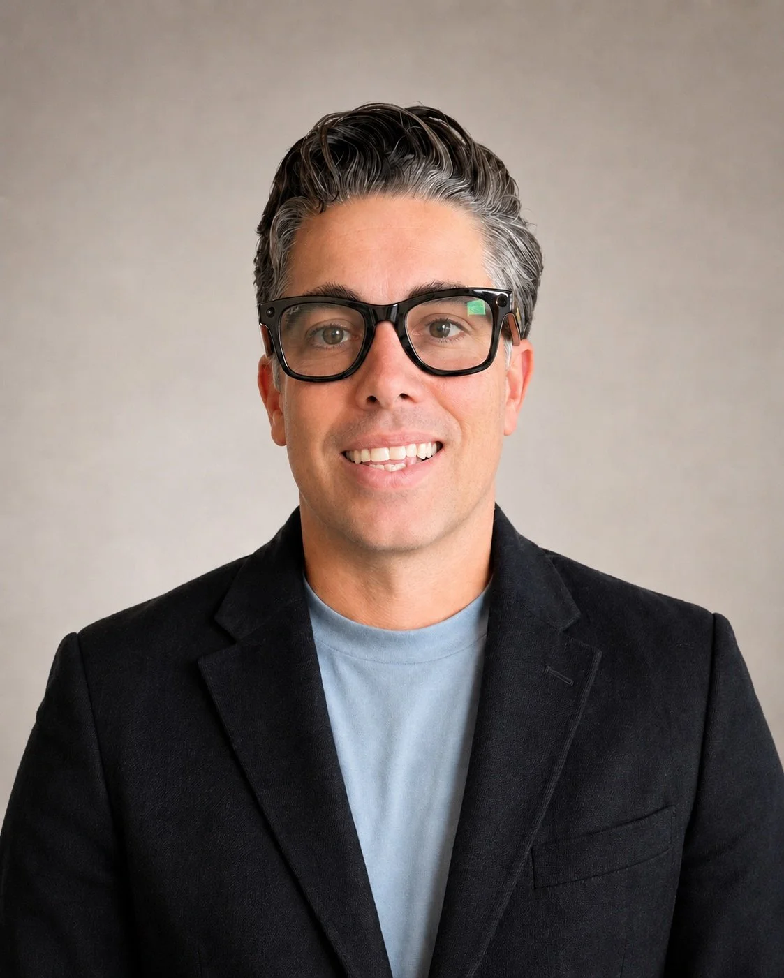 A man with dark, slicked-back hair, wearing large black glasses, a light blue shirt, and a black blazer, smiling at the camera against a plain gray background.