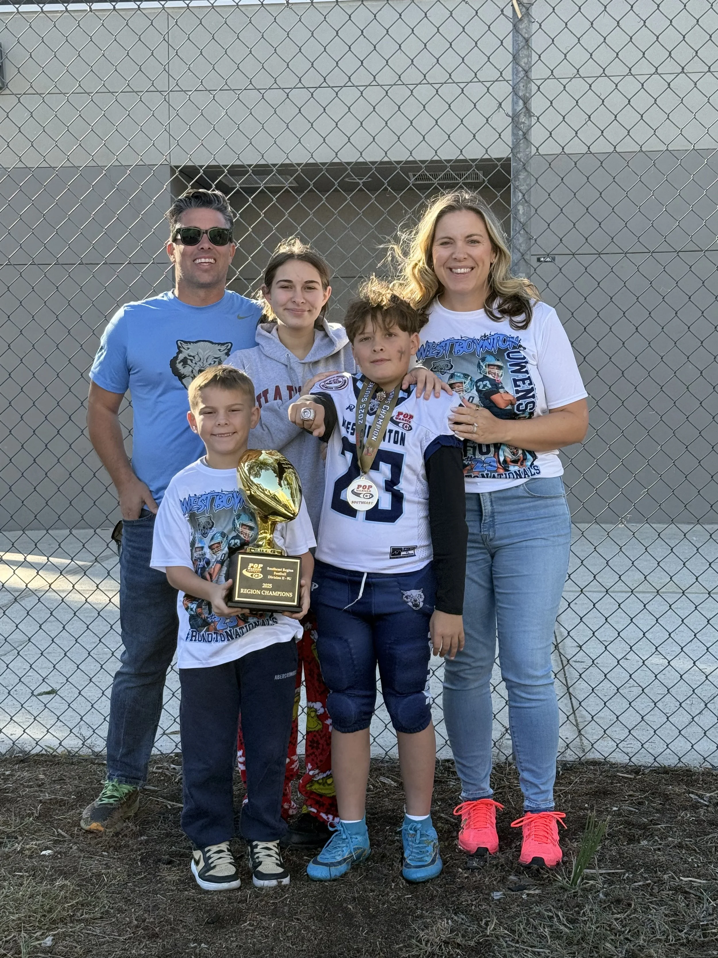 Group of five people standing outdoors in front of a chain-link fence, with the sun setting in the background. Two children in the front, one holding a trophy and wearing a sports jersey with a medal around his neck. Three adults behind, smiling and standing close together.