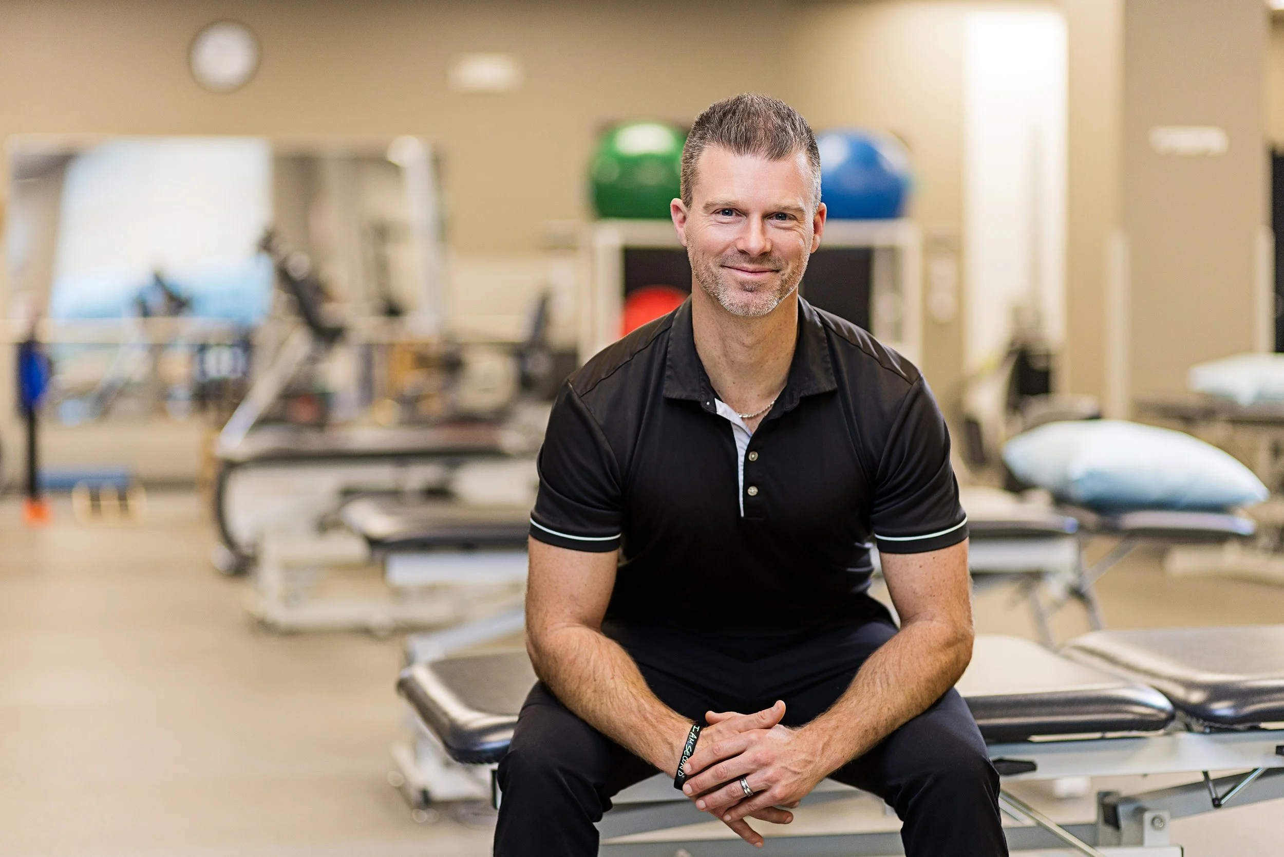 Physical therapist in a black polo seated in a Pilates studio with reformer machines, Indianapolis branding portrait