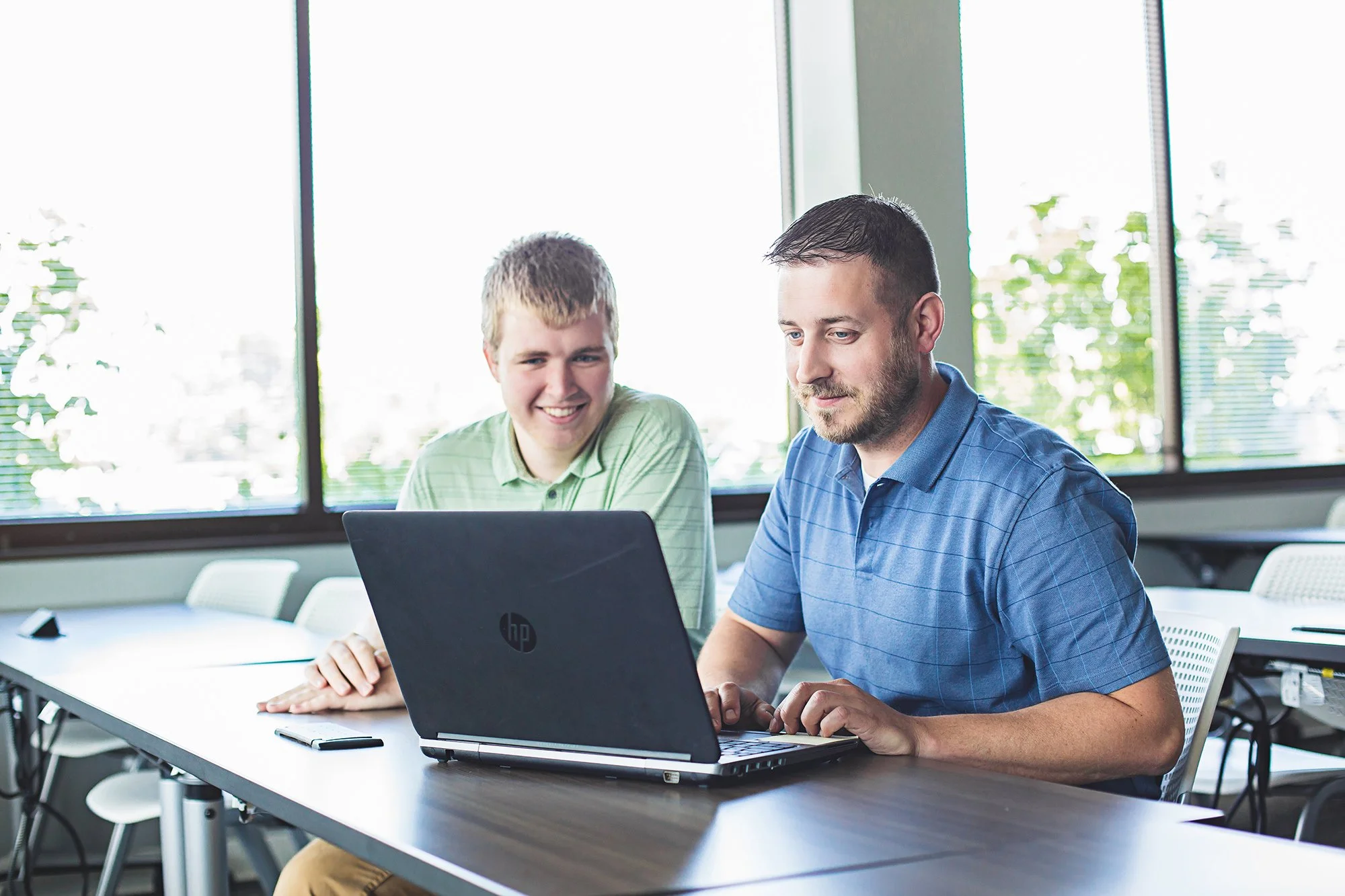 Two colleagues working on laptops at a table by large windows, BCforward office brand photography