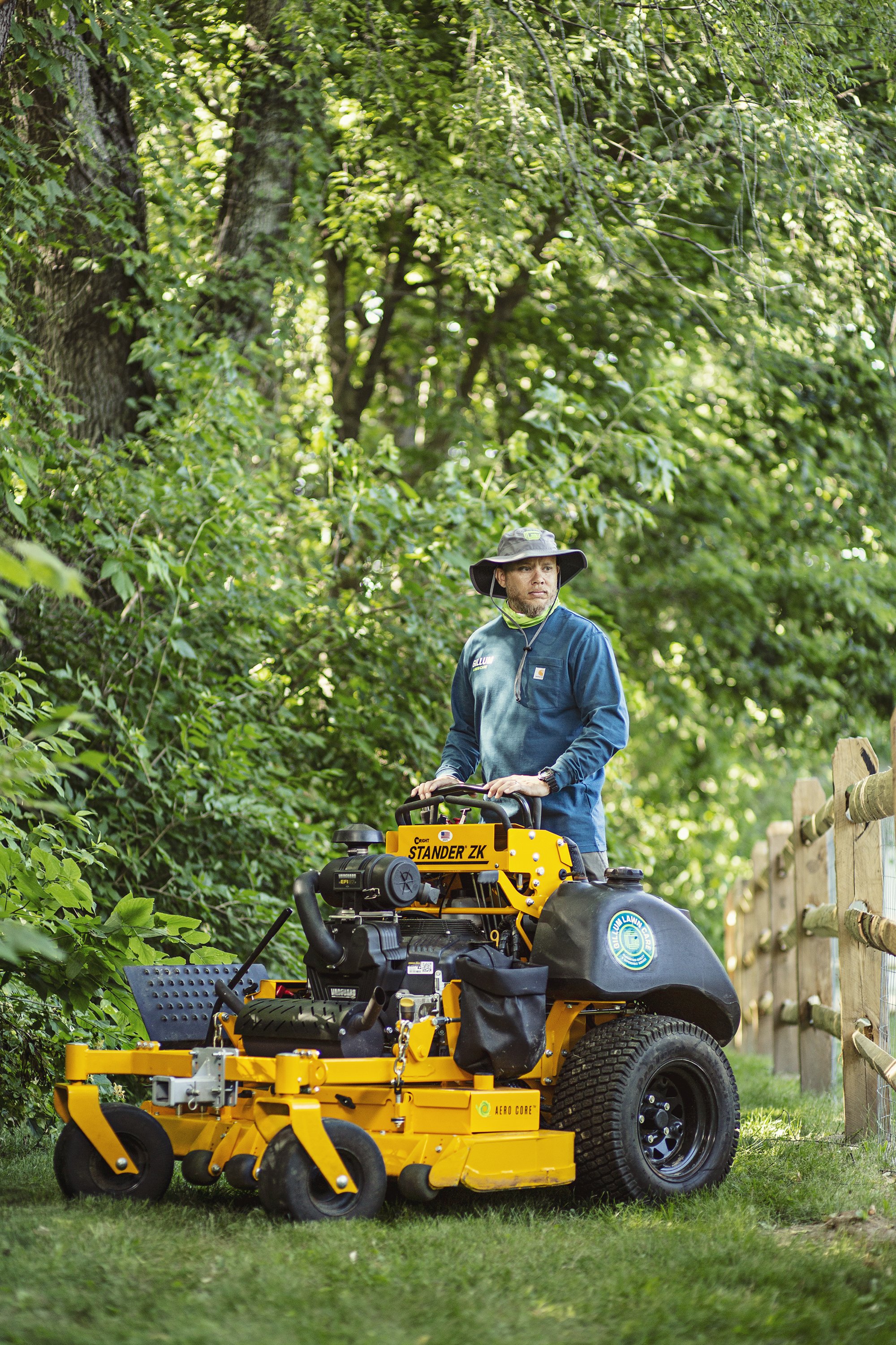 Lawn care professional on a yellow stand-on mower beside a wooded yard, Indianapolis brand photography