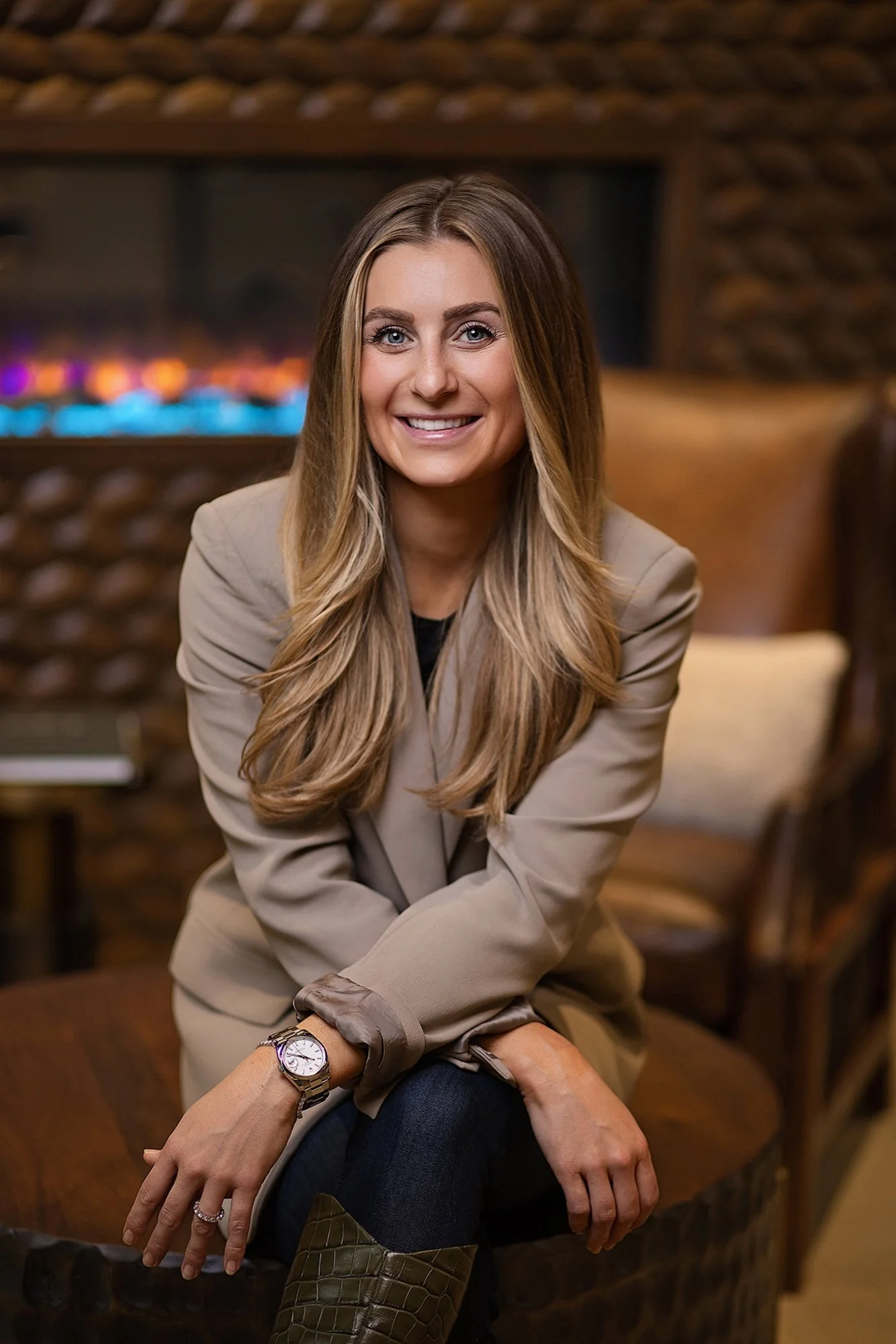 Smiling woman in a tan blazer seated by a fireplace, warm-toned branding portrait by Square Mouth Studios