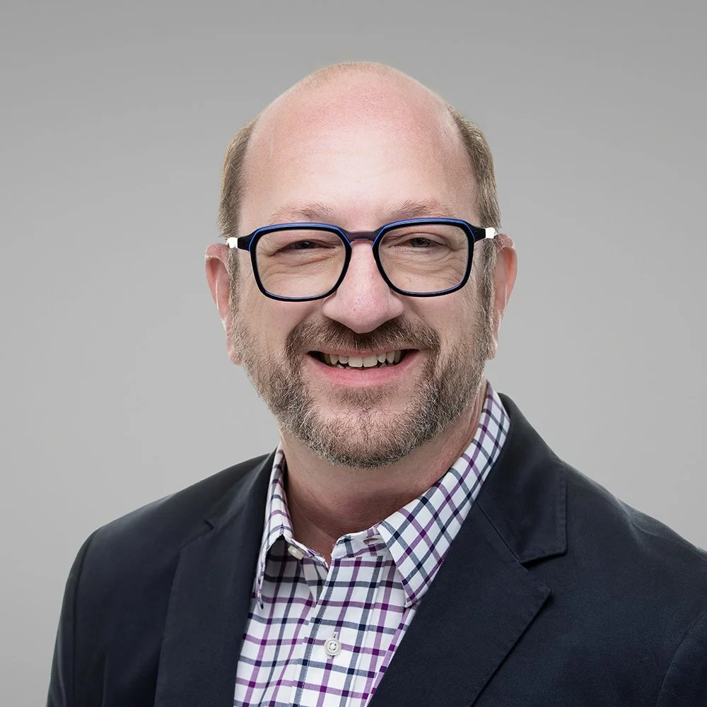 Tom Darling smiling in glasses, a navy blazer and checked shirt, professional headshot